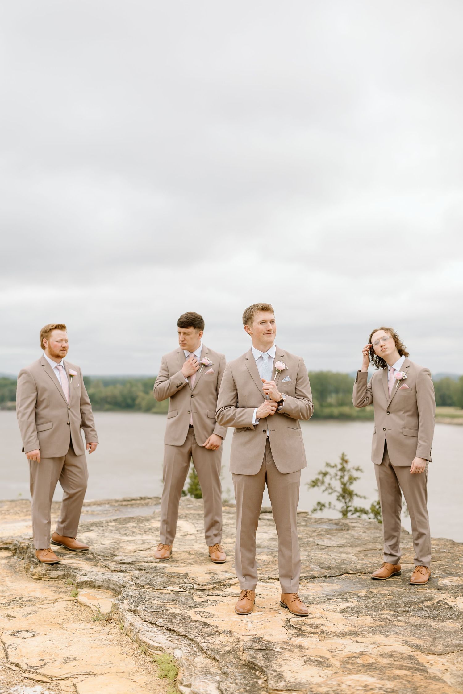 Groomsmen in tan suits stand along a rocky shoreline overlooking a lake on an overcast day.