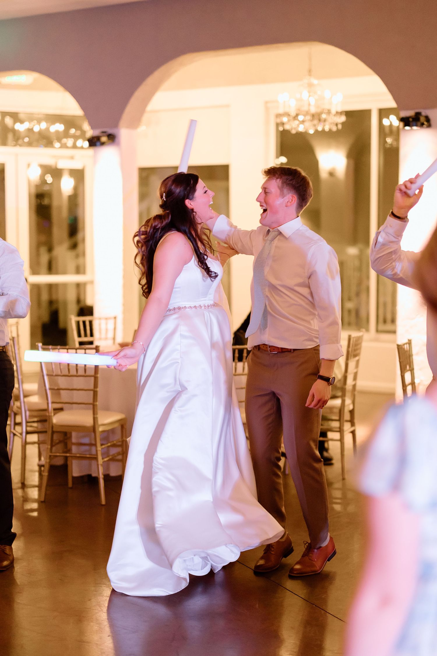 Guests dancing at an elegant wedding reception in a bright white ballroom with arched doorways and chandeliers.