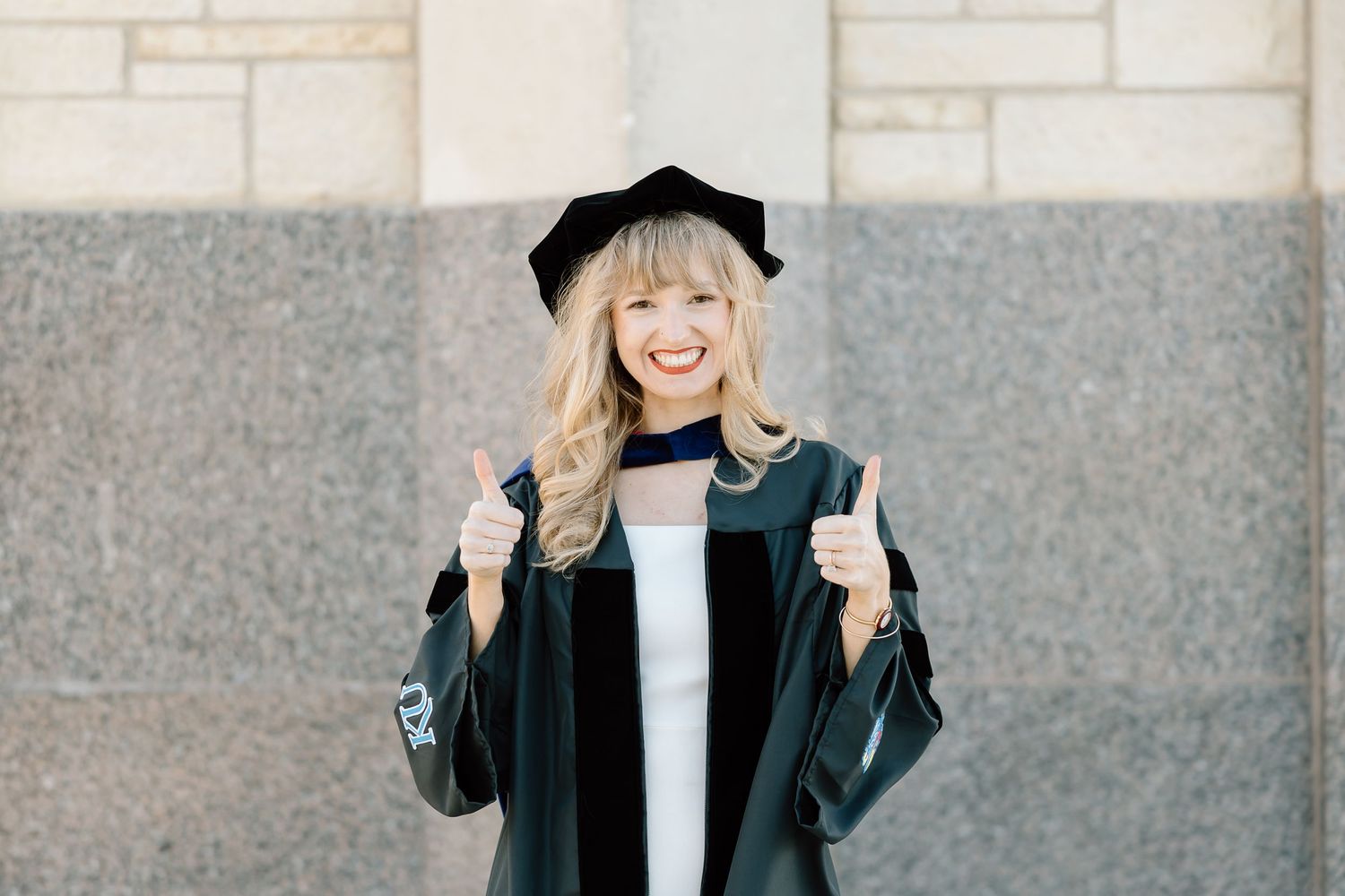 Graduate in cap and gown gives thumbs up while smiling against stone wall background.