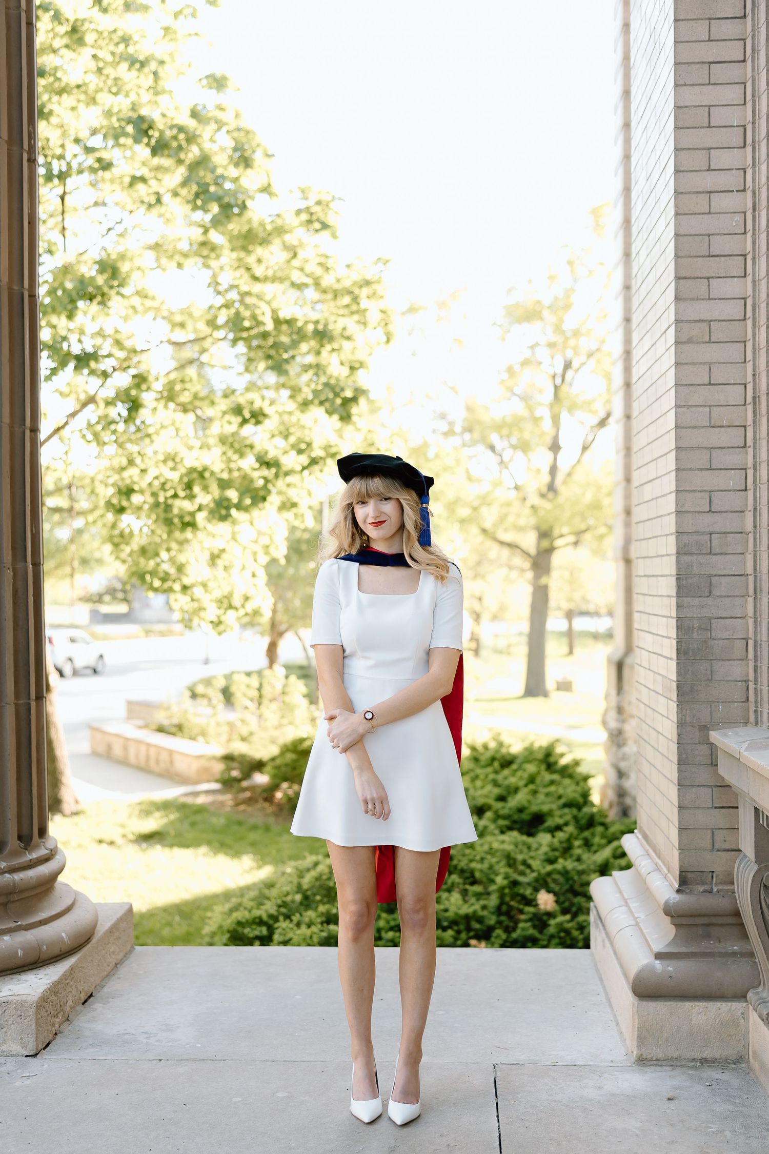 A person in white dress and shoes stands gracefully between architectural columns on a sunny day.