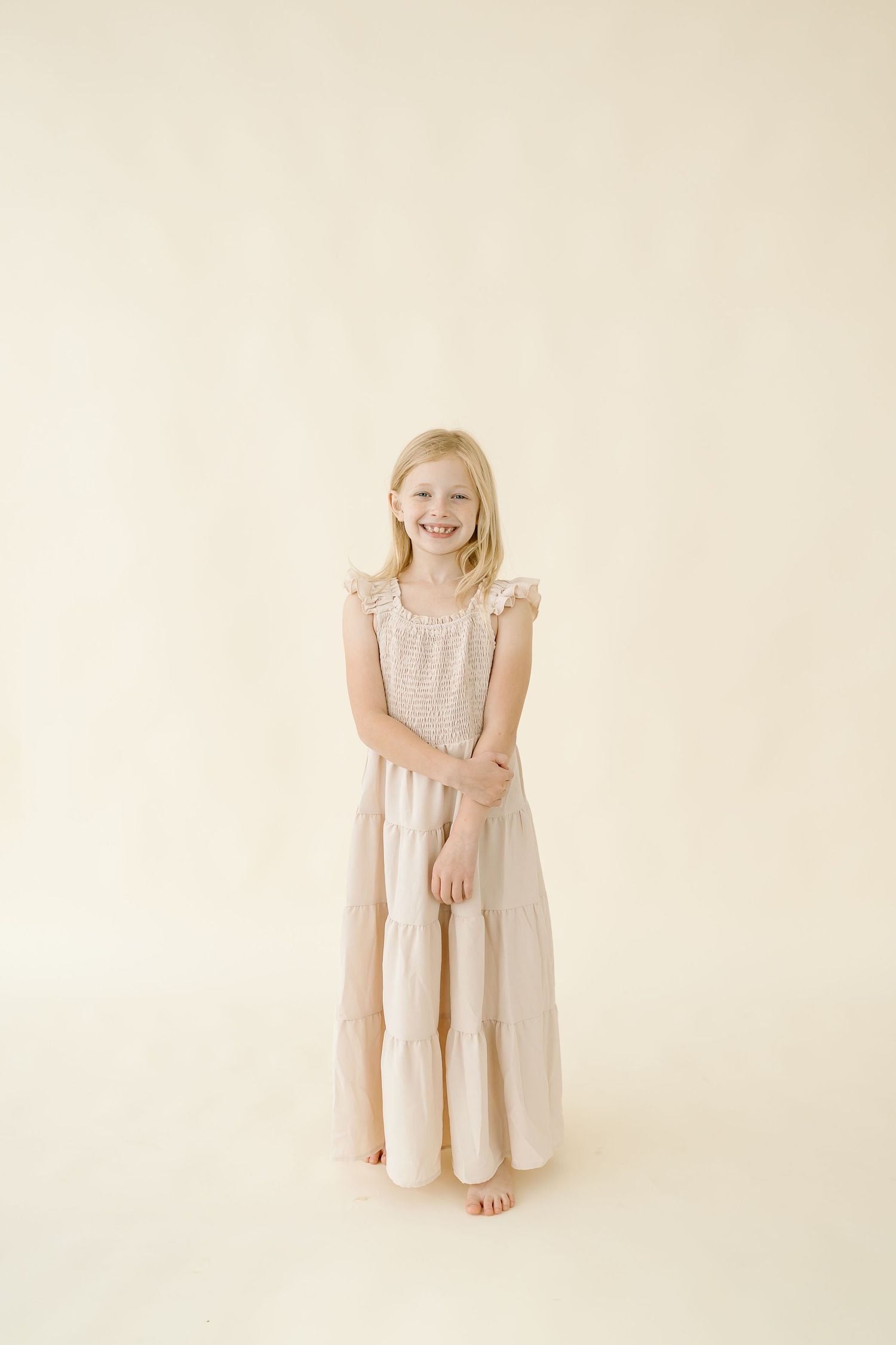 A barefoot figure in a flowing cream-colored tiered maxi dress stands against a light beige background.