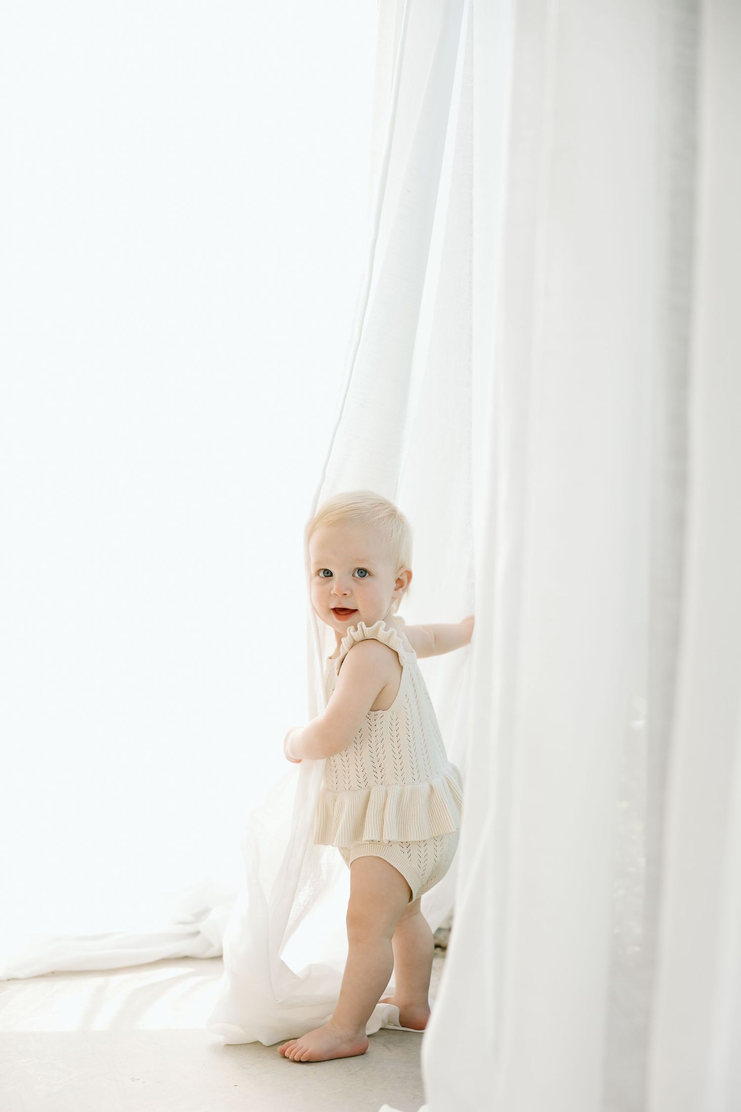 Baby in white knit outfit crawls on floor near sheer curtains in bright natural light.