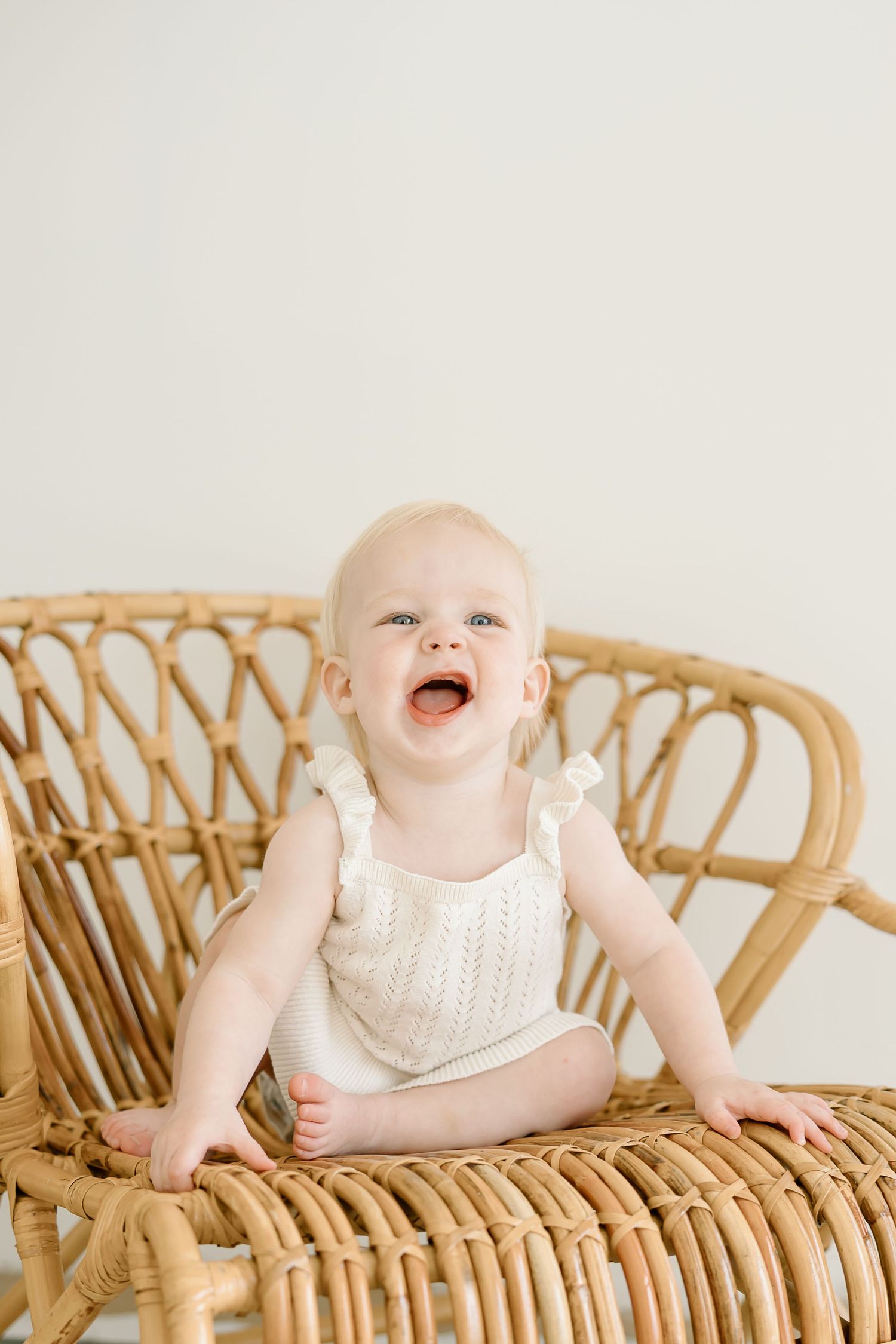 Baby in white sleeveless outfit sits and smiles while posing on vintage rattan furniture against light background.