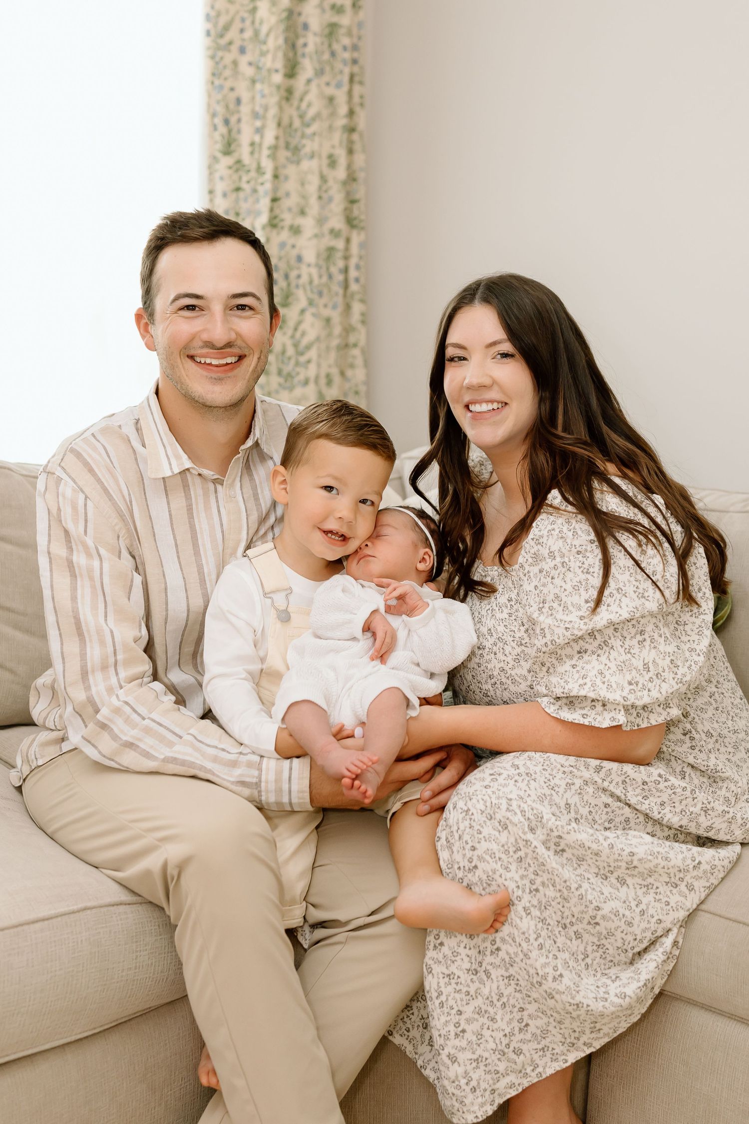 A family sits together on a beige couch in a bright room with floral curtains.