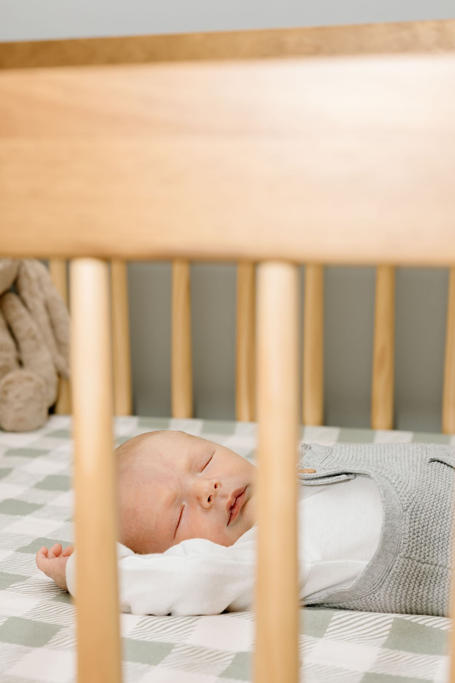 A sleeping baby lies peacefully in a wooden crib with grey bedding visible through the slats.