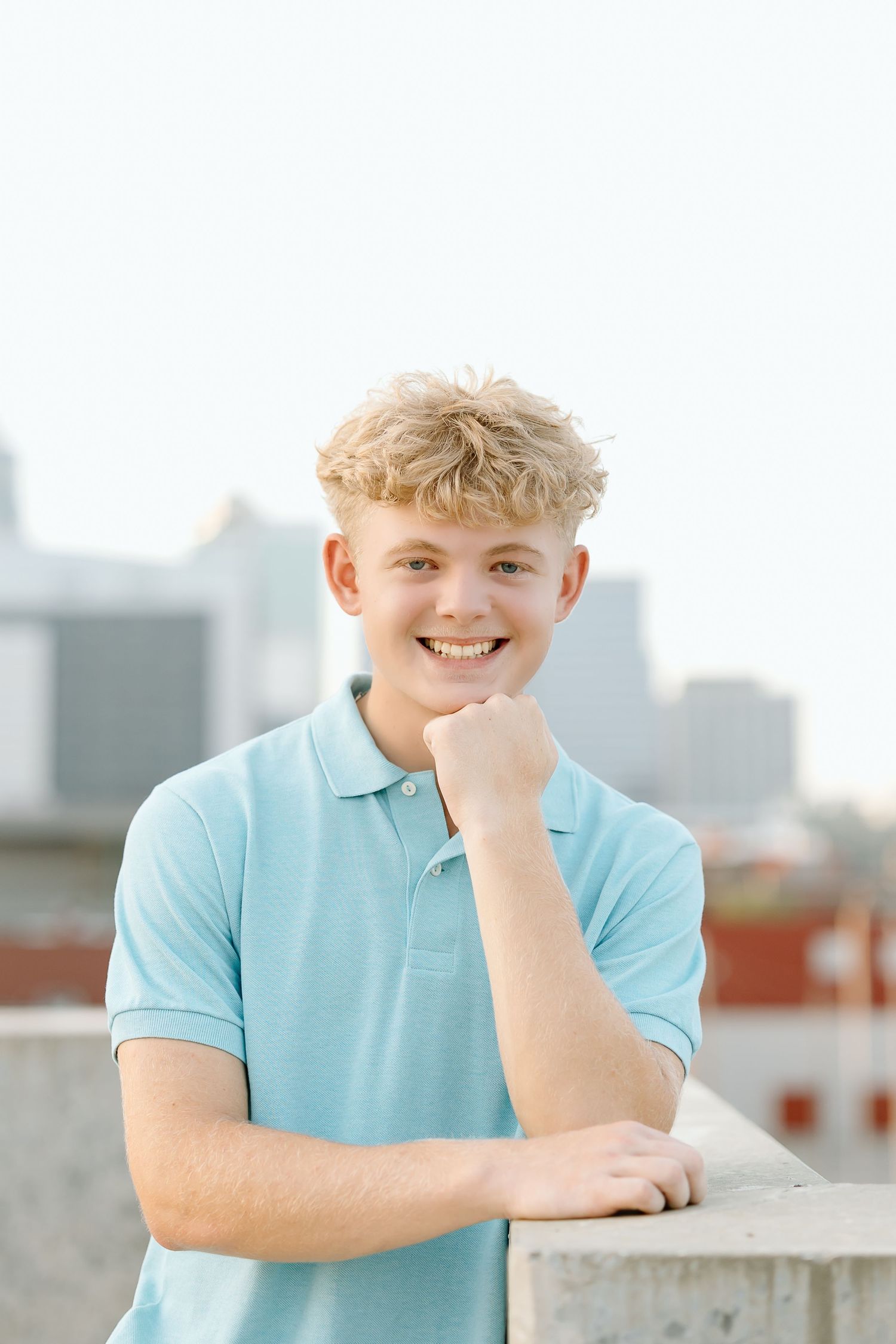 A person wearing a light blue polo shirt poses casually against an urban rooftop backdrop.