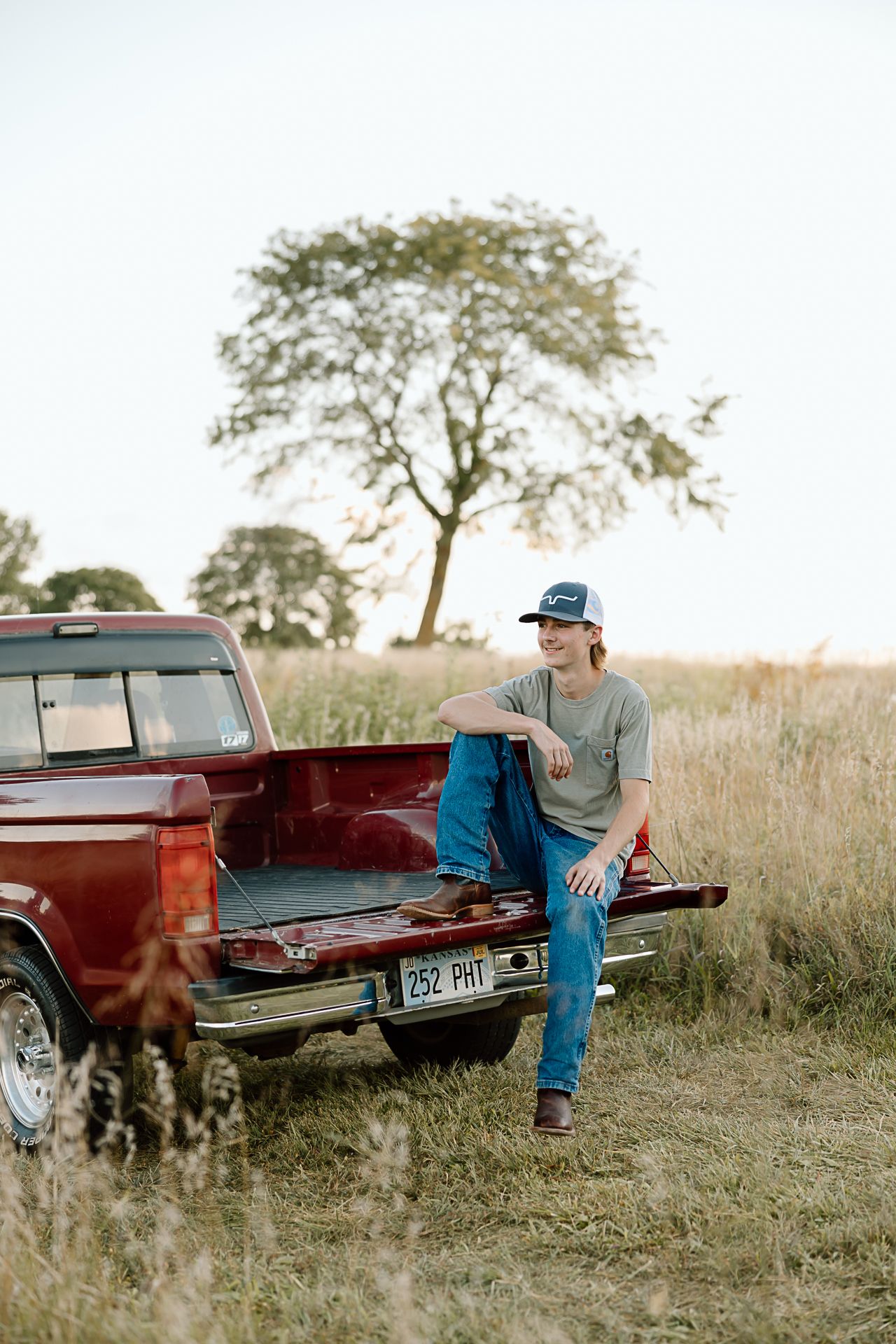 A rustic scene of someone sitting in the bed of a red pickup truck in a grassy field at sunset.