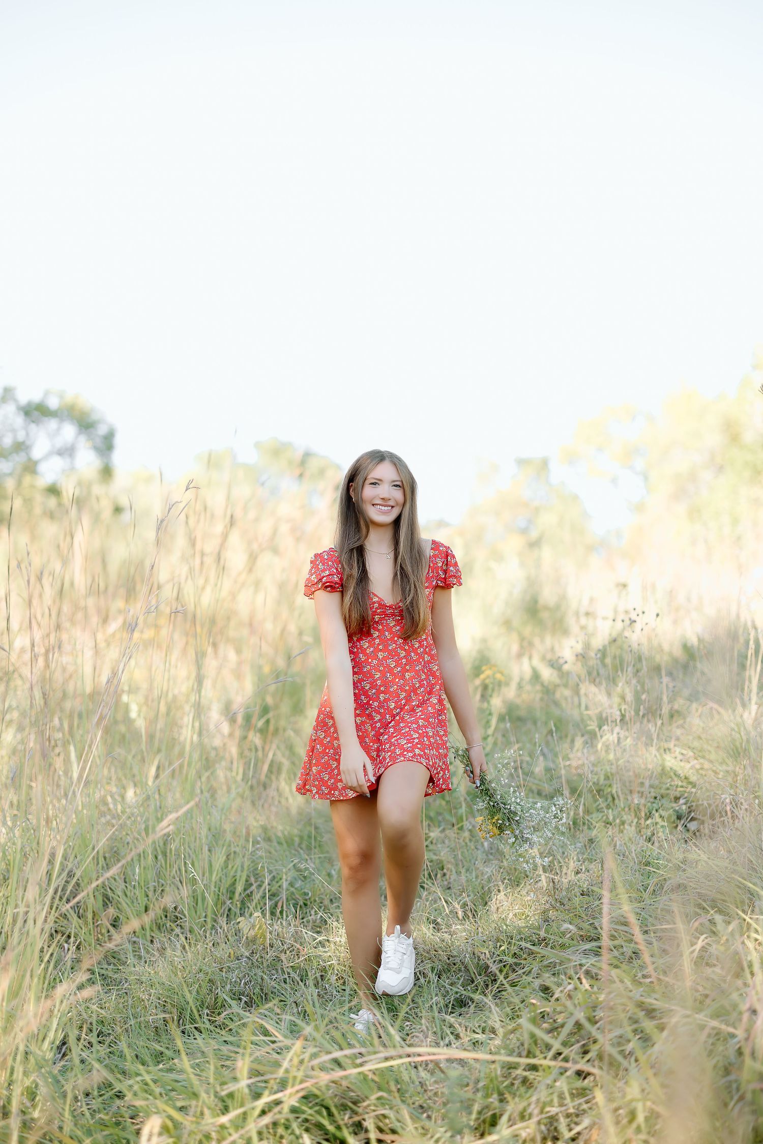 A person wearing a red floral dress walks through a sunlit meadow of tall grass during golden hour.