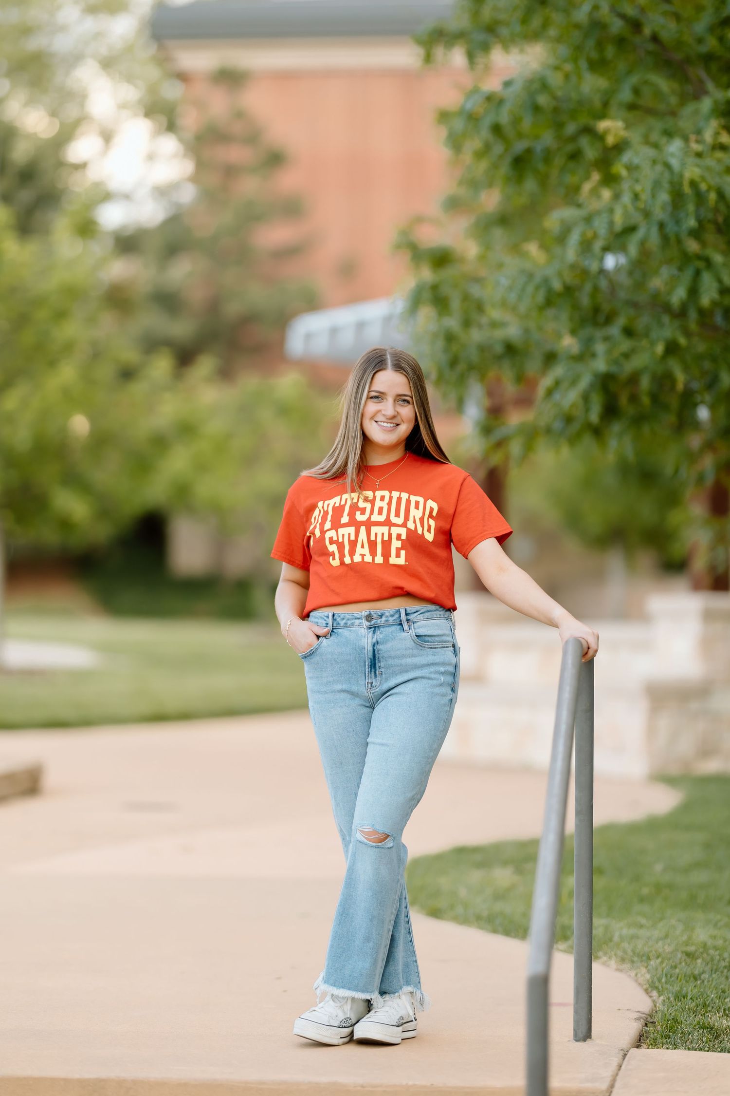Person in orange university shirt and ripped jeans poses casually on campus walkway in outdoor photoshoot series.
