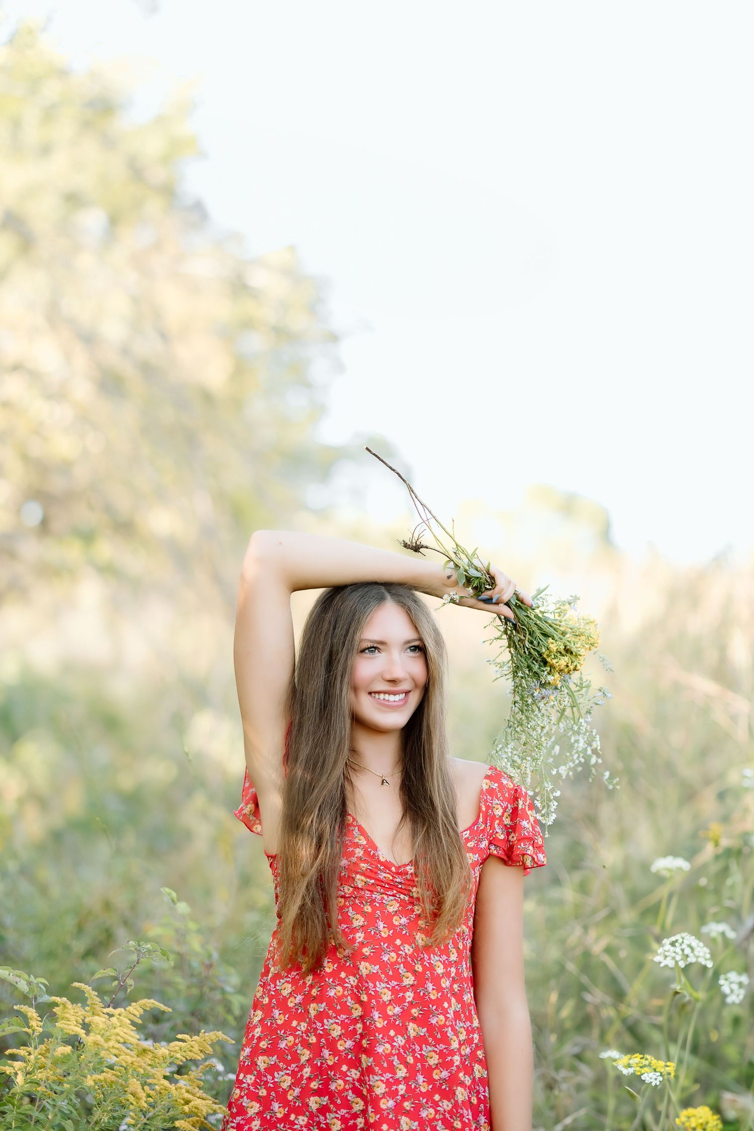Person in a red floral dress poses playfully with wildflowers in a sunny meadow during golden hour.