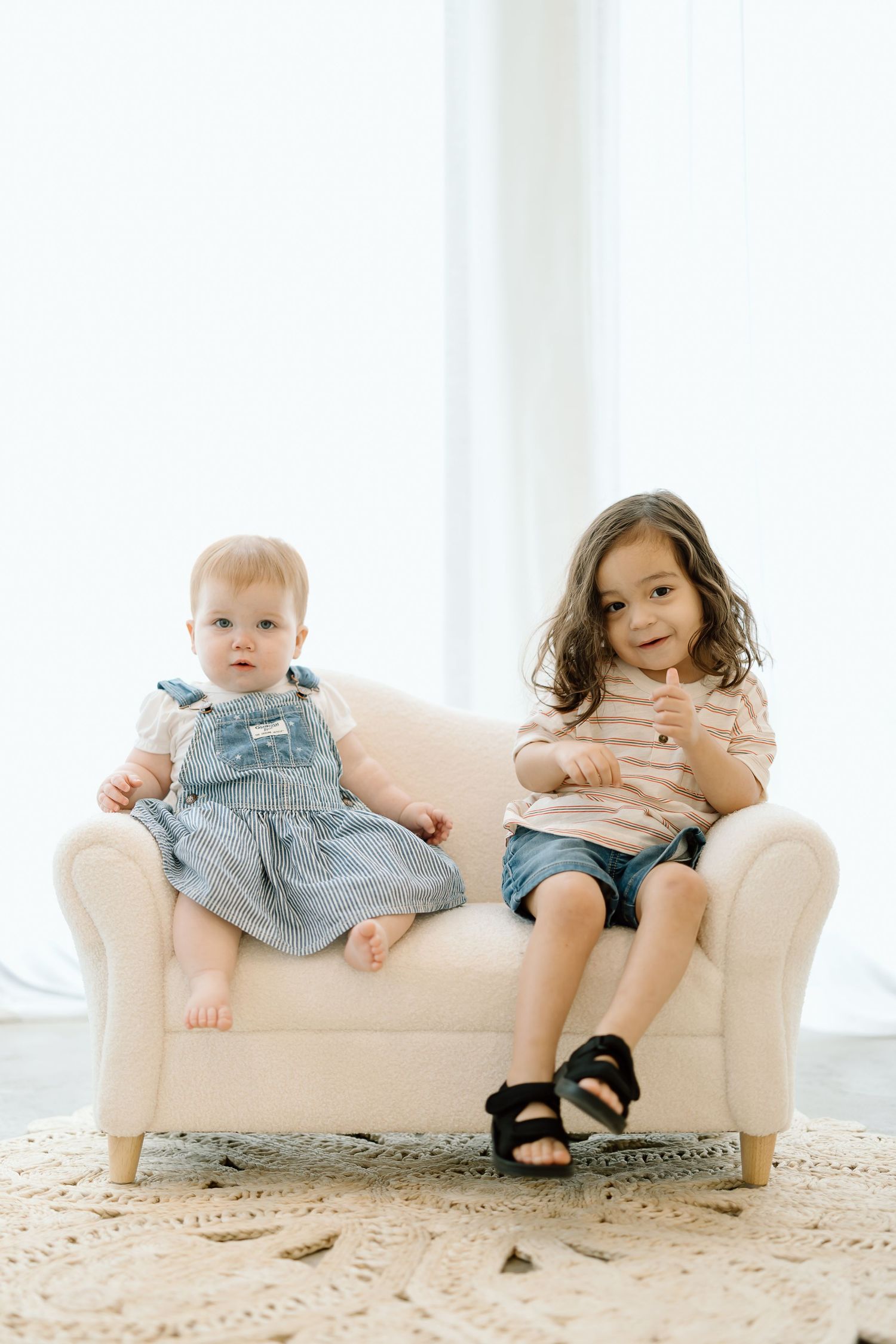 Two children sitting together on a small cream-colored couch against a white wall with natural lighting.