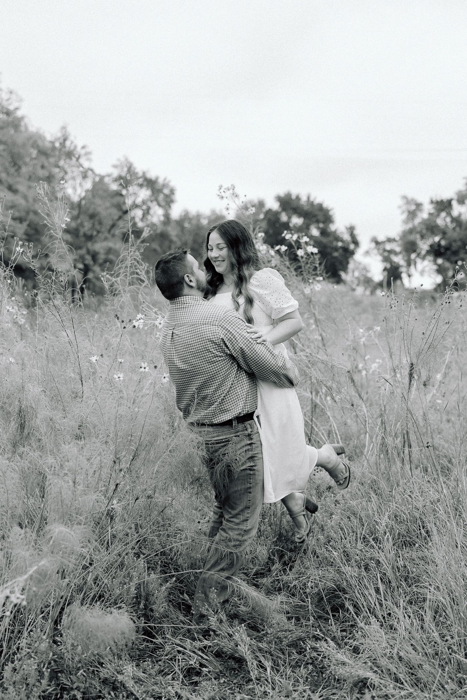 A black and white photo captures a couple's romantic lift pose in a wild grass field.