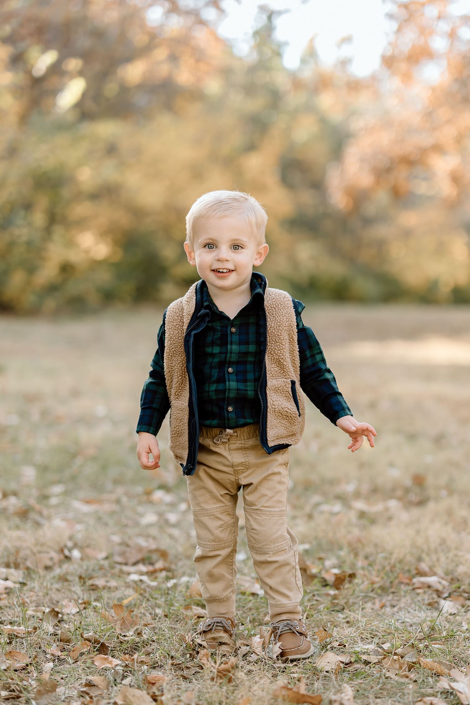 Smiling toddler in plaid shirt and brown vest stands in fallen leaves during autumn.
