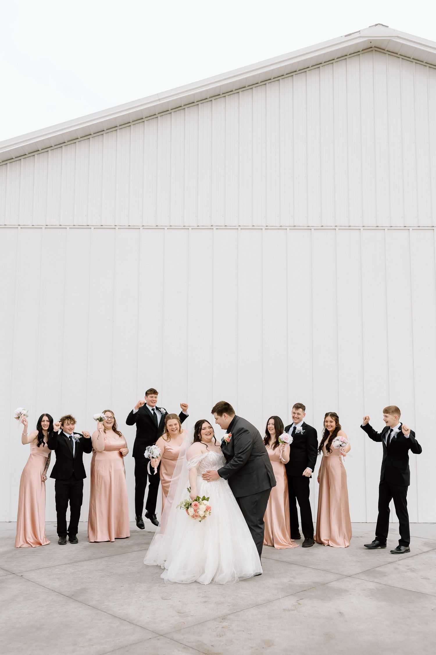 Wedding party poses joyfully in front of white barn with bridesmaids in blush dresses.