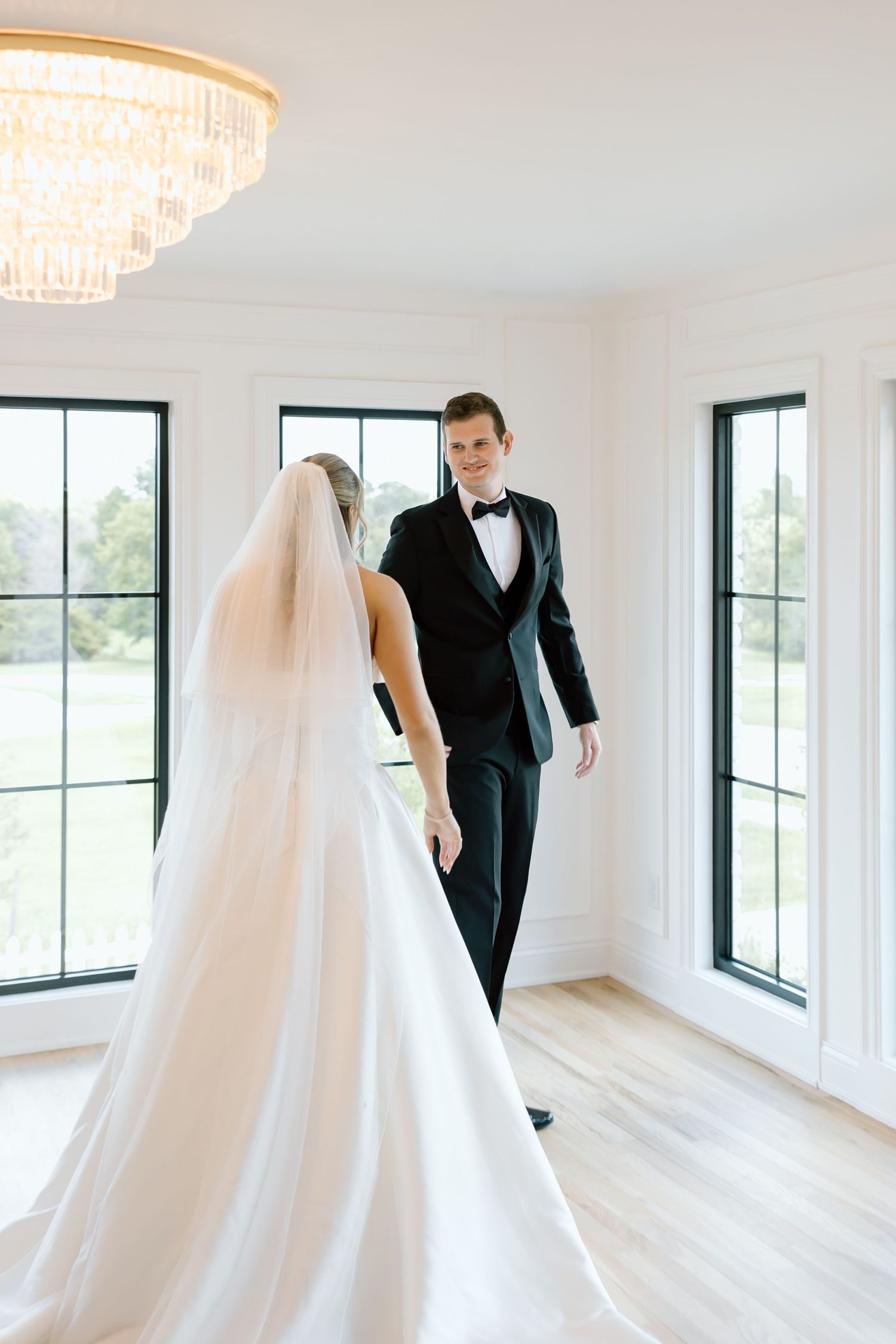 Bride and groom share a first look moment in a bright white room with large windows and an elegant chandelier.