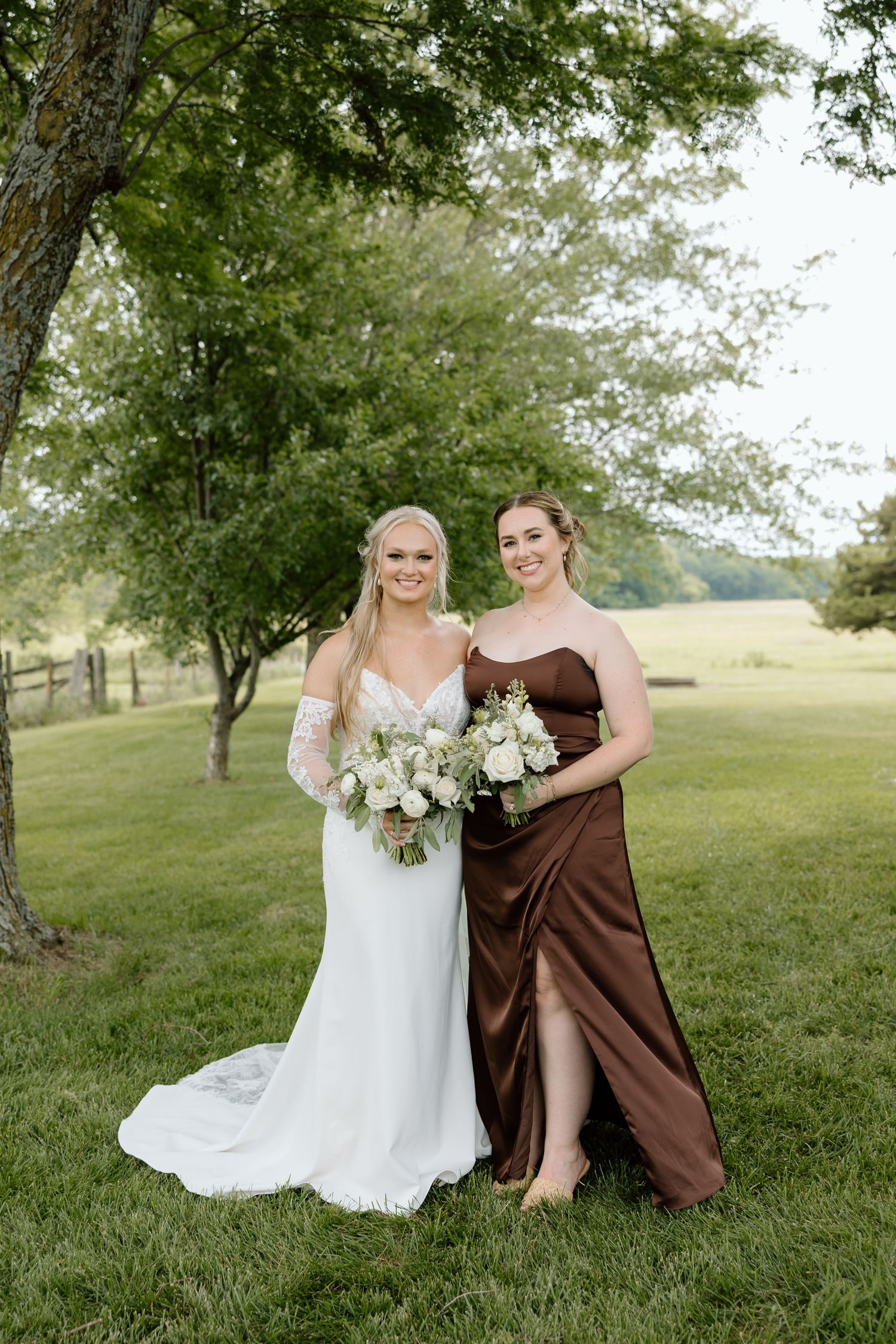 Bride in white lace gown and bridesmaid in brown satin dress holding white bouquets outdoors on green lawn.