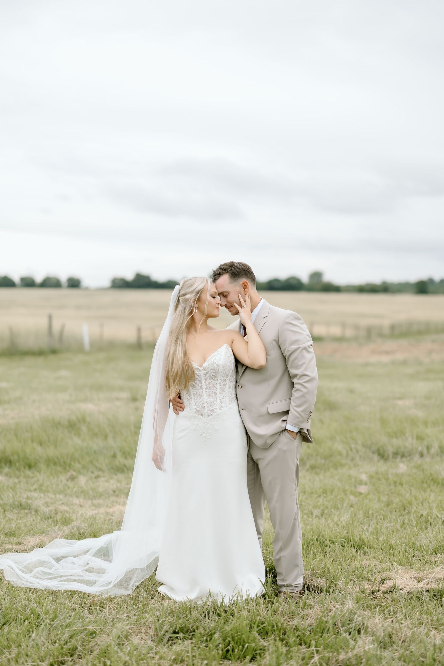 A bride and groom share a romantic moment in a lush green field, with her white gown and veil trailing behind.