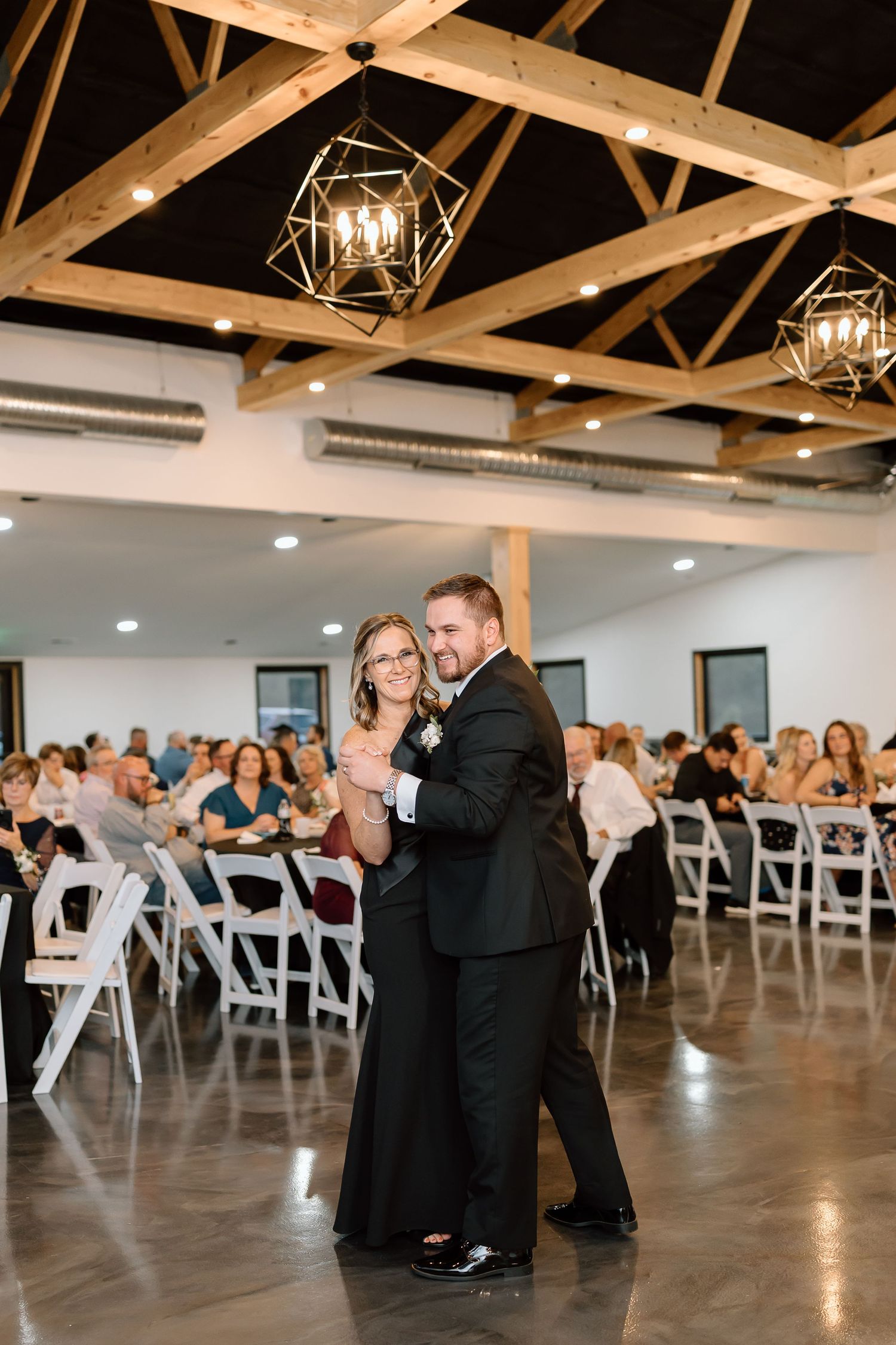 A couple dances together in an elegant venue with wooden beam ceilings and white folding chairs for guests.