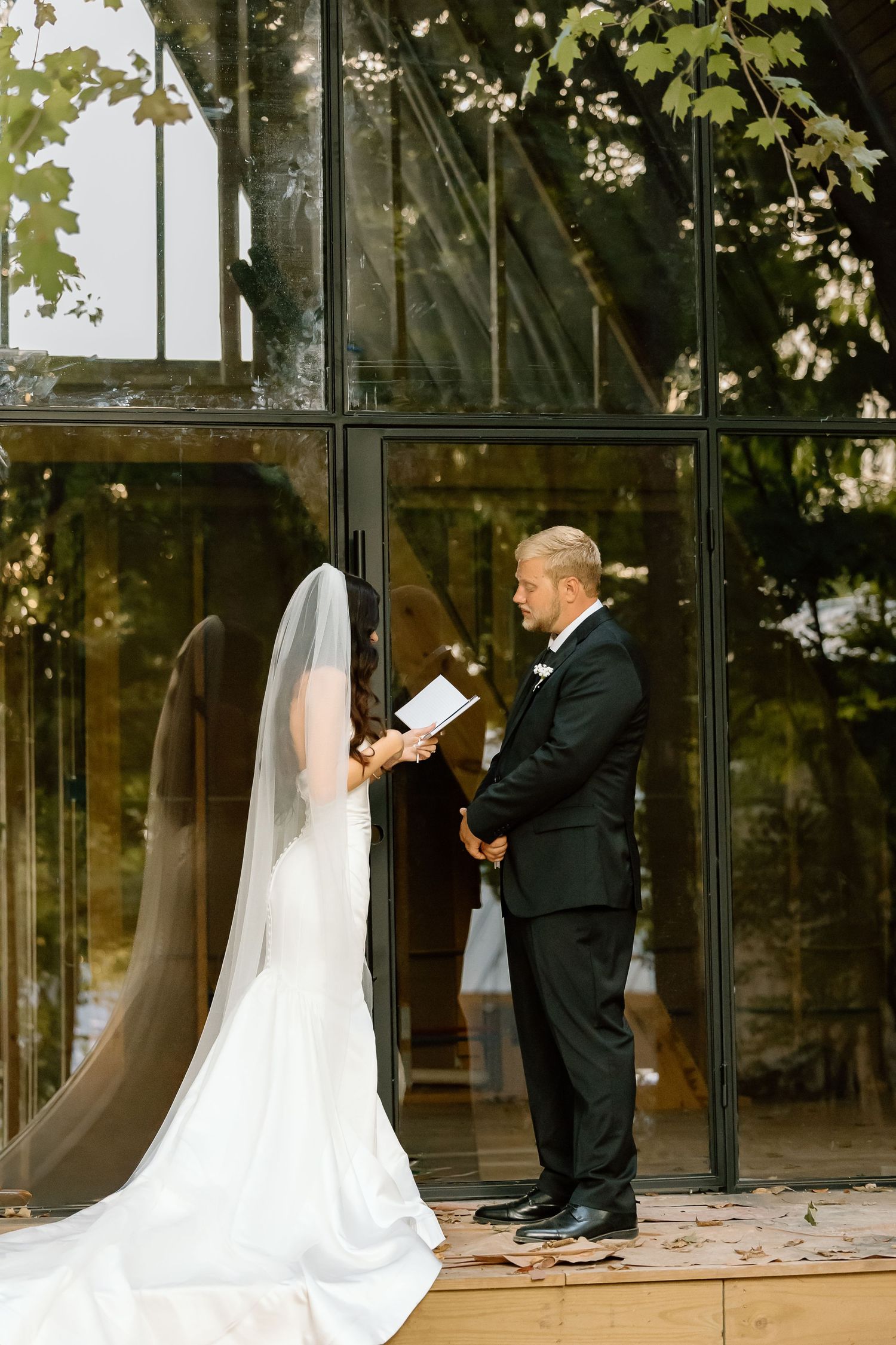 A bride and groom share a tender moment during their first look outside a glass-walled building.