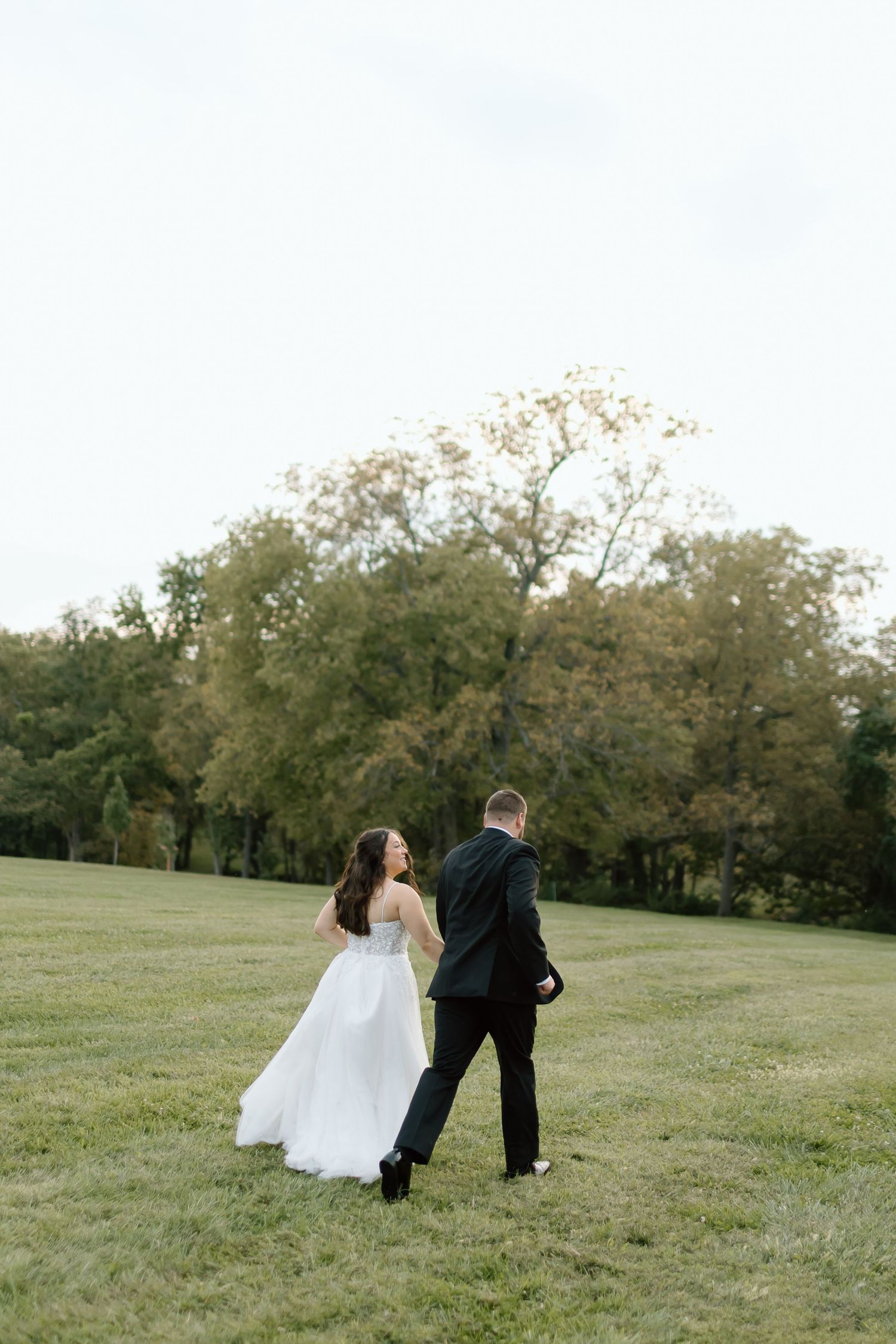 Newlyweds walk playfully together across a grassy hillside on their wedding day.