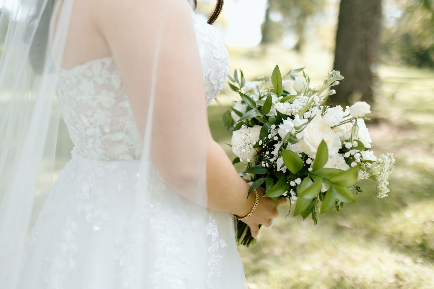 A bride holds a delicate white and green floral bouquet against a soft, sunlit outdoor background.