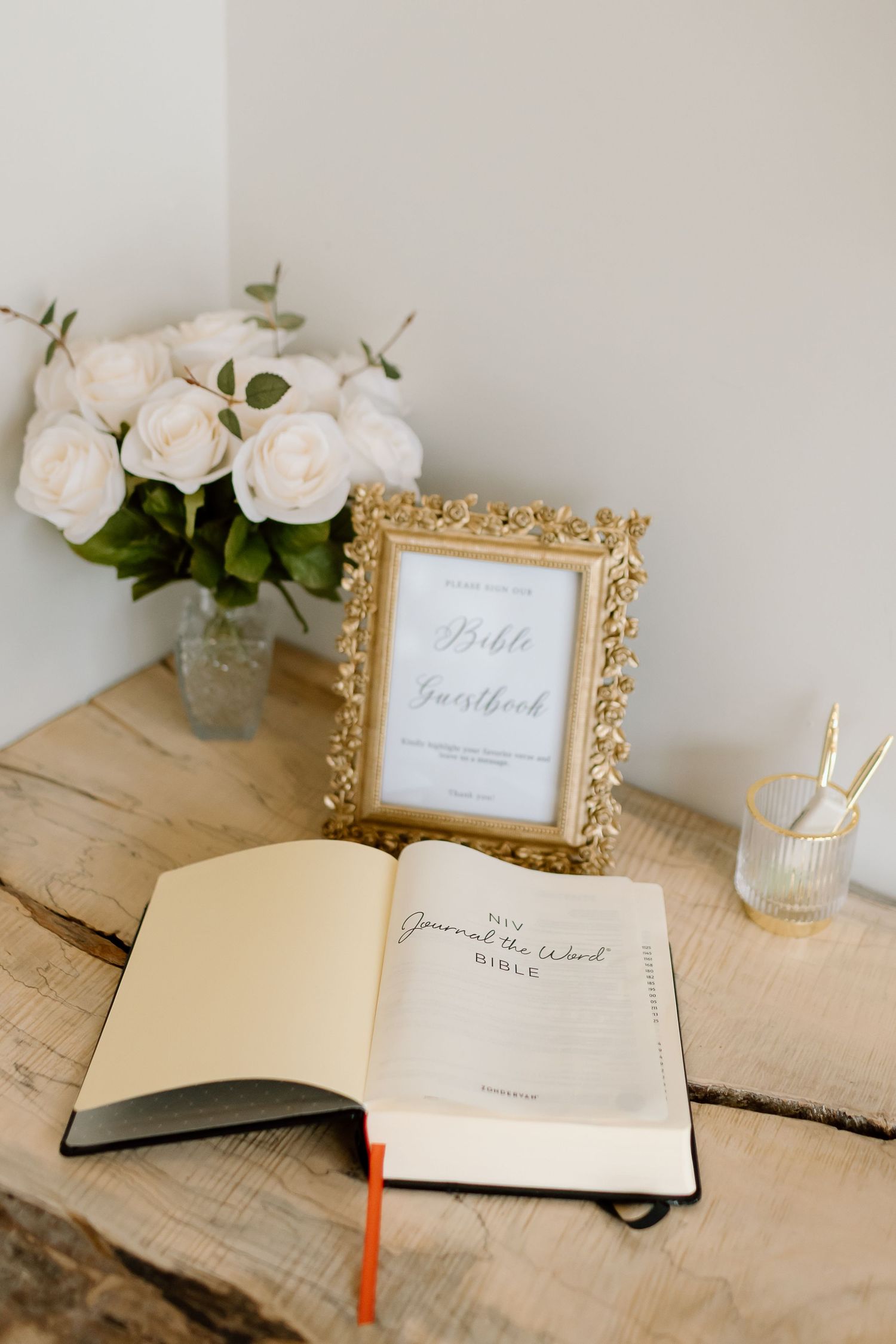 A rustic wedding guestbook setup with white roses and gold frame on a wooden table with elegant decor details.