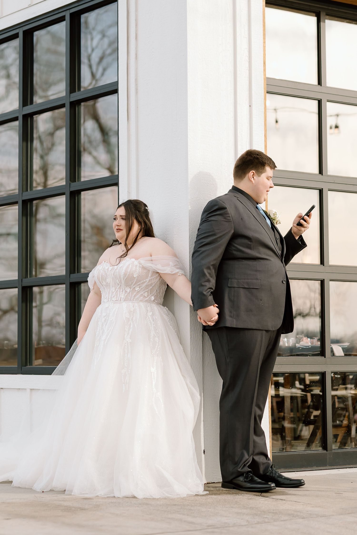 A bride in a flowing white gown and groom in dark suit stand apart looking at their phones in a modern venue.
