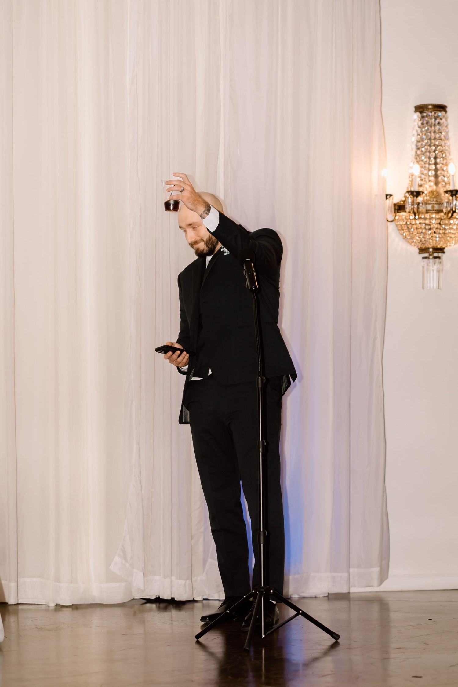A person in a black suit checks their phone while standing next to a microphone stand and crystal chandelier.