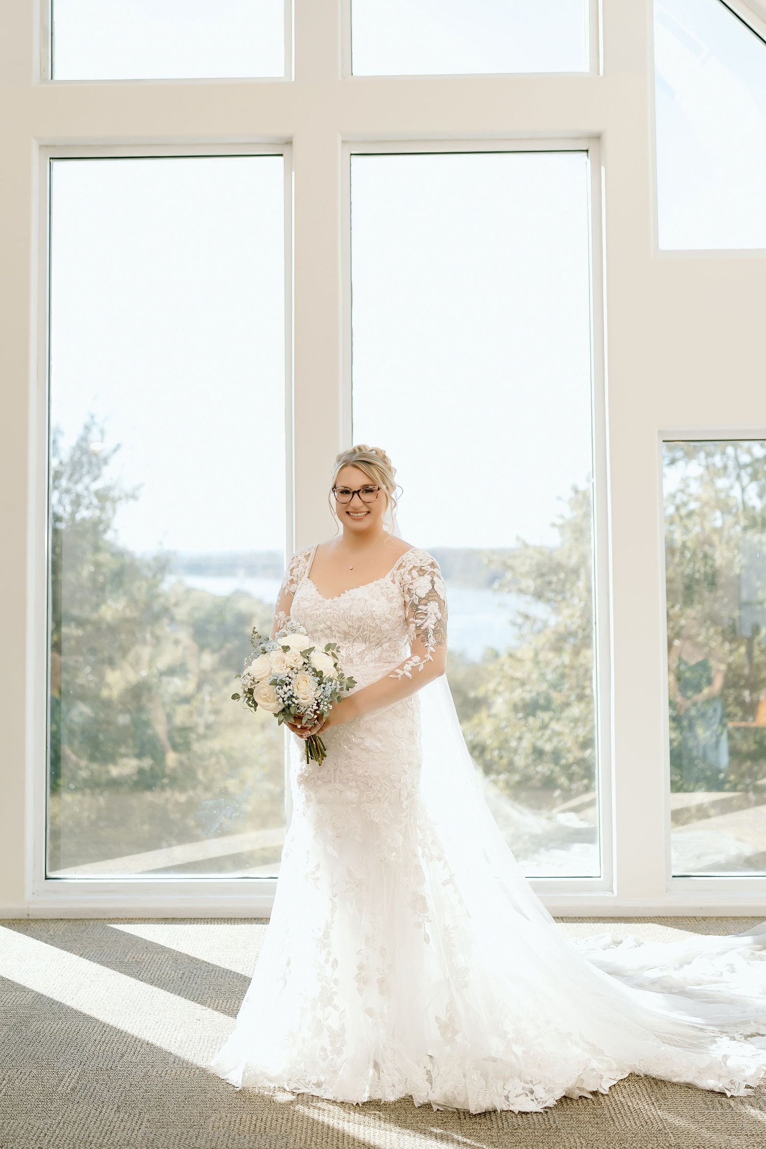 A bride in a flowing lace wedding gown stands by large windows with natural light streaming in.