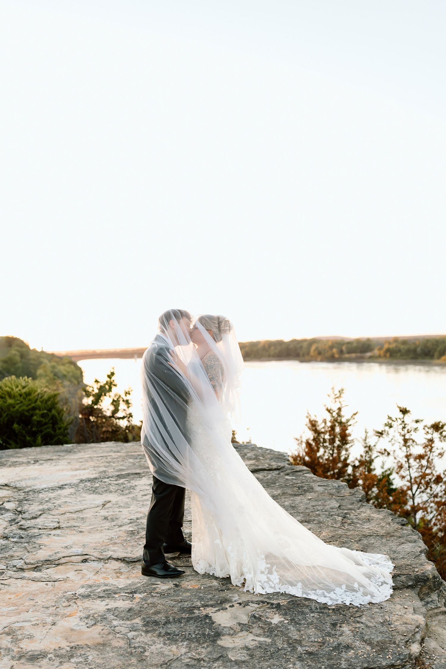 A romantic couple in wedding attire share an intimate moment on a rocky cliff overlooking a serene lake at sunset.