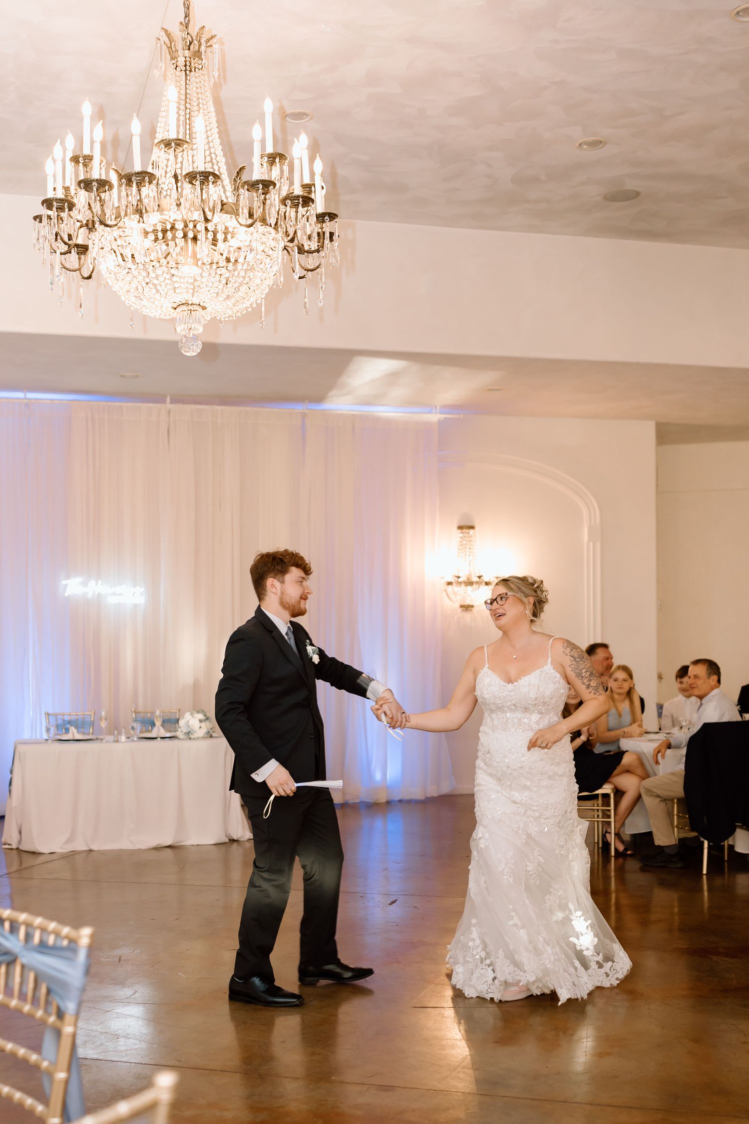 Couple performs a choreographed dance routine in an elegant ballroom with crystal chandeliers and white decor.