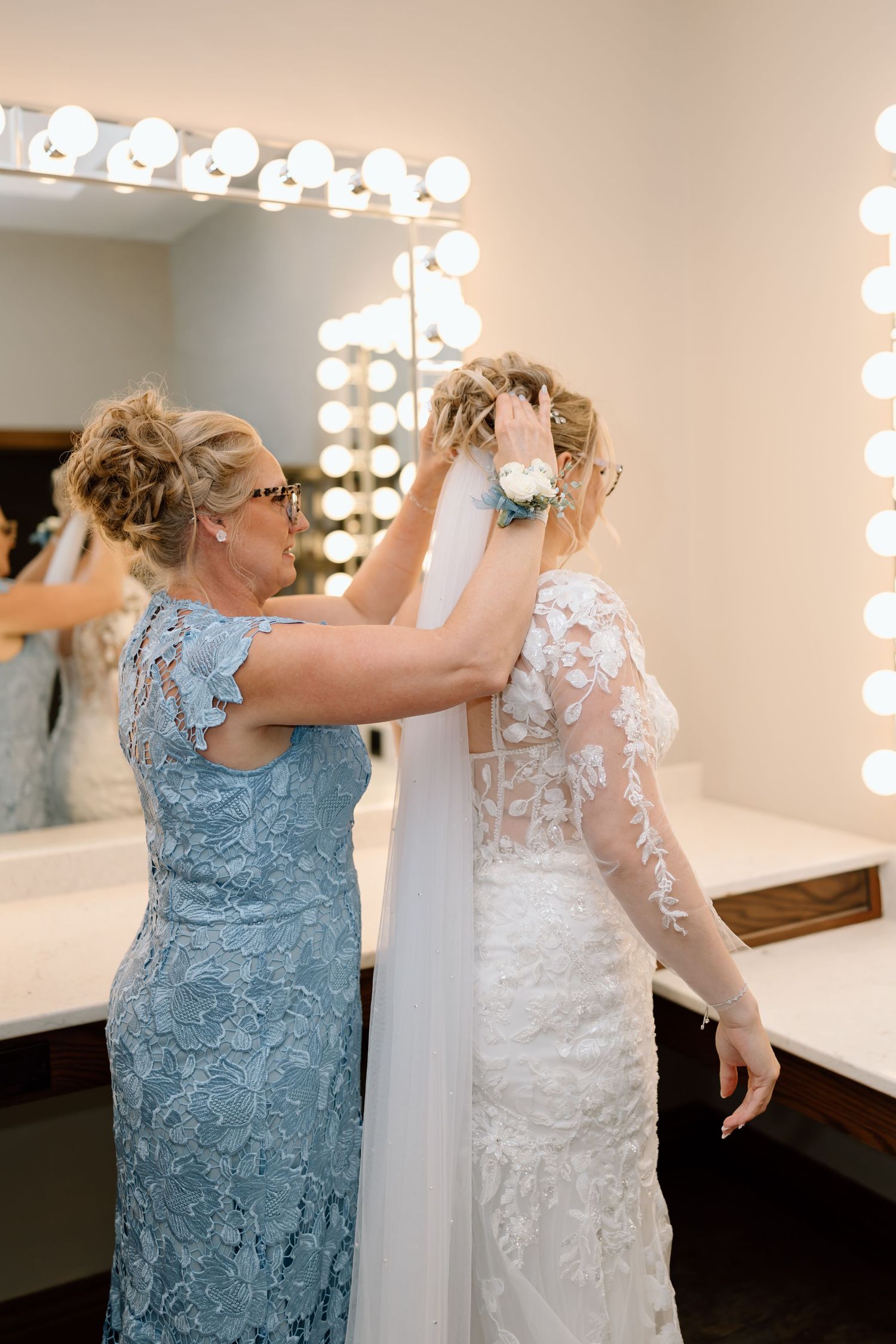 A woman in a blue lace dress hangs a white wedding gown on a wall in a dressing room with illuminated mirrors.