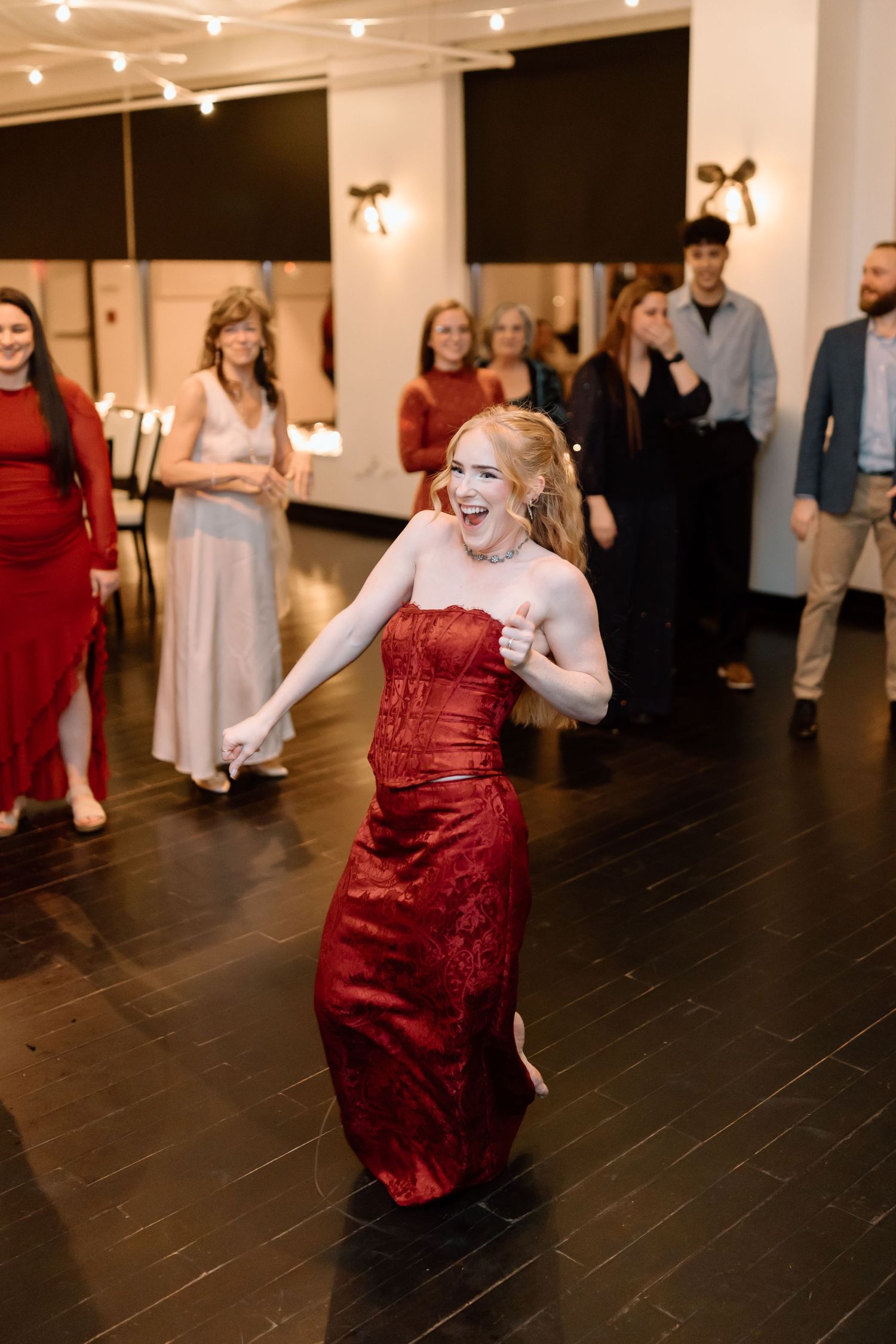 A person in a stunning red formal dress dances joyfully on a dark wooden dance floor at an indoor event.