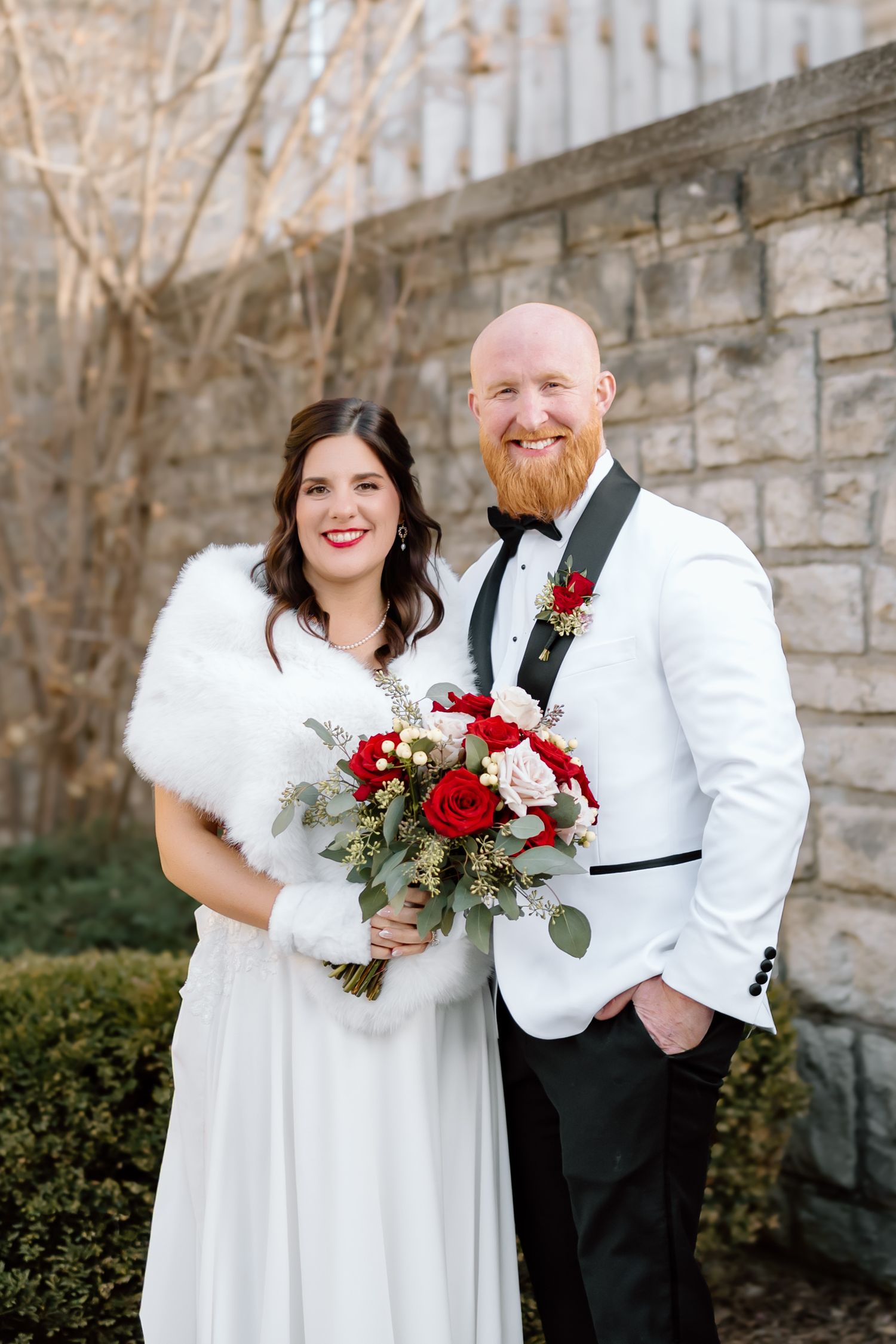 Wedding couple in winter attire share joyful moments against stone wall backdrop with red rose bouquet.