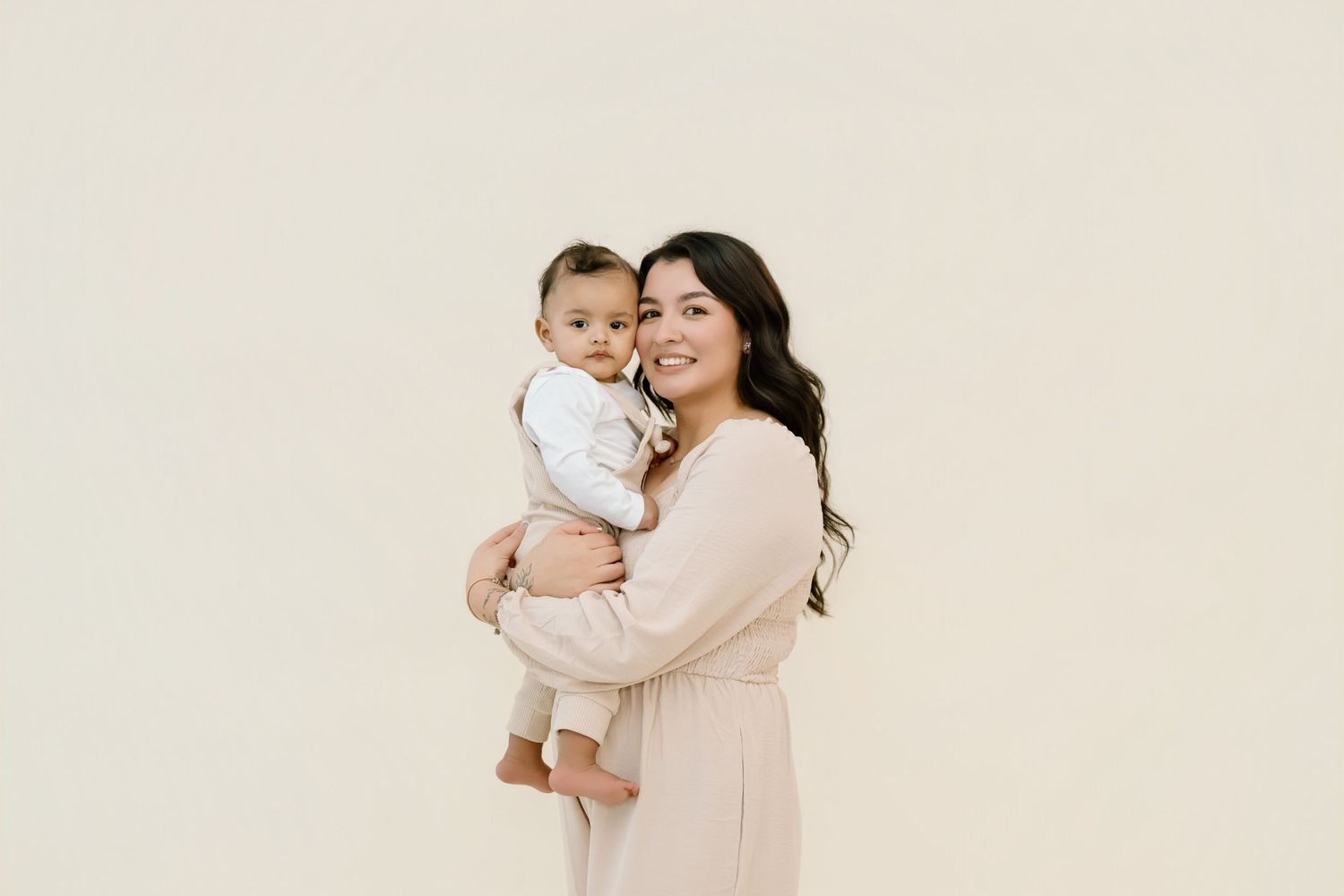 A parent in a cream sweater holds and embraces a baby in matching light-colored outfits against a neutral background.