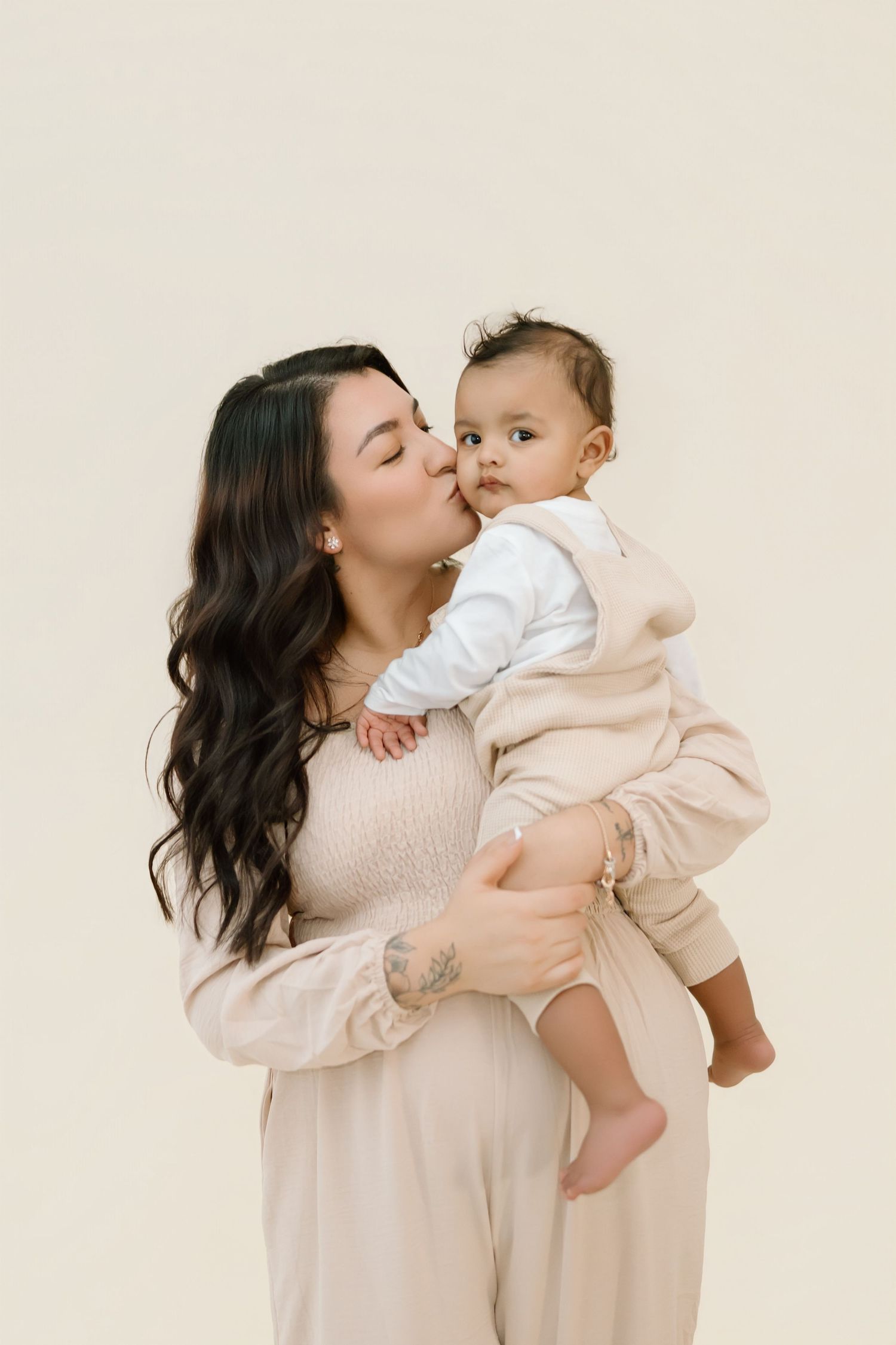 Mother and baby in matching cream outfits share tender moments in minimalist studio portraits.