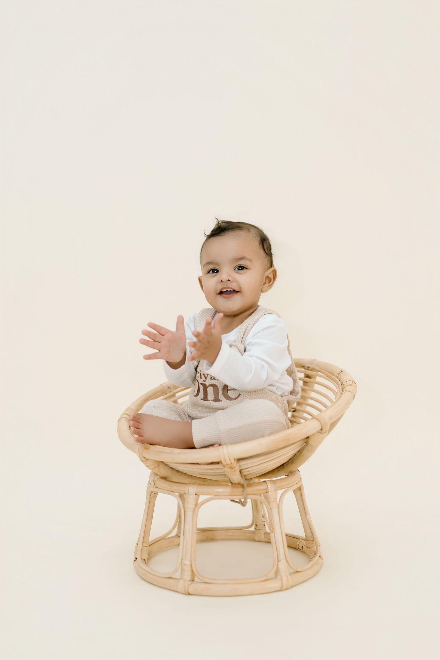 Adorable baby in white onesie sitting and smiling in natural wooden rocking chair against neutral background.