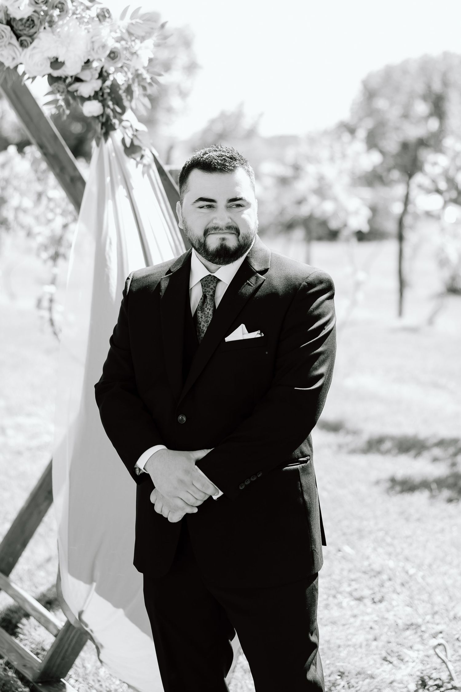 Smiling groom in a classic black tuxedo poses outdoors for wedding photos against a light background.