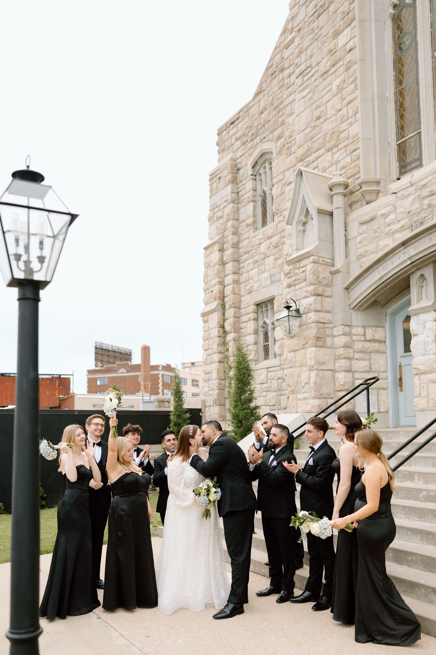 Wedding party in black attire celebrates outside a stone church with bridesmaids holding white bouquets.