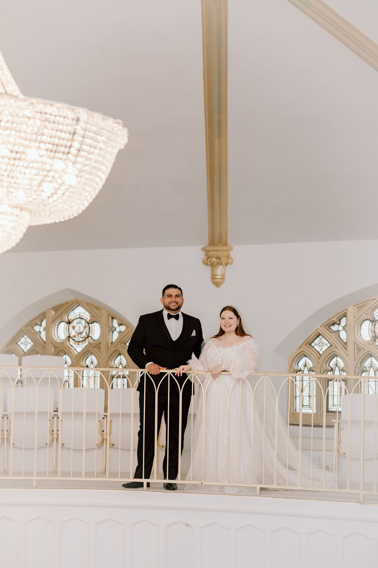 Bride and groom standing on elegant chapel balcony with arched stained glass windows and crystal chandelier.
