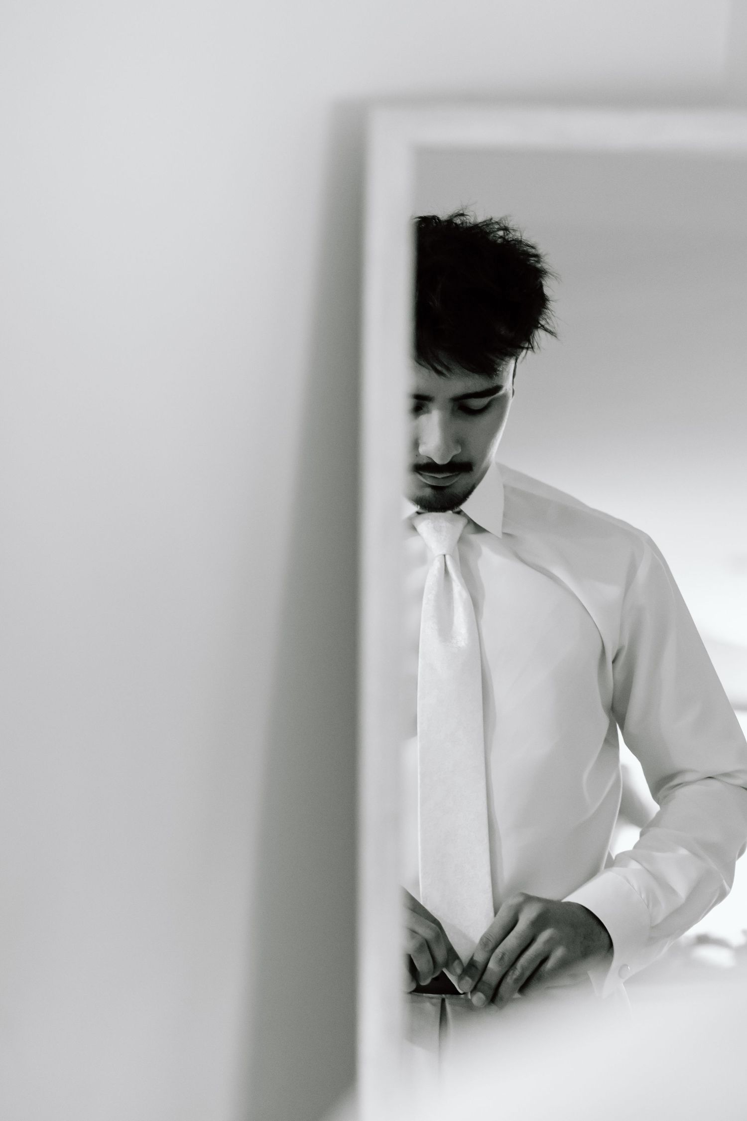 Black and white photo of a groom buttoning his shirt while wearing a white tie, reflected in a mirror.