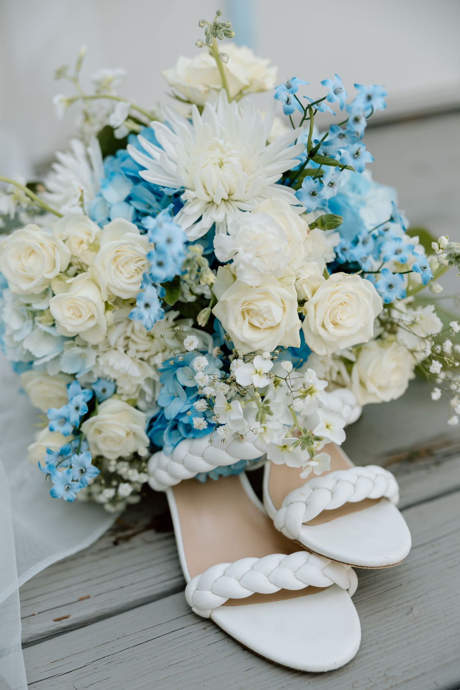 White braided bridal heels rest in front of a lush wedding bouquet with white roses and blue flowers.
