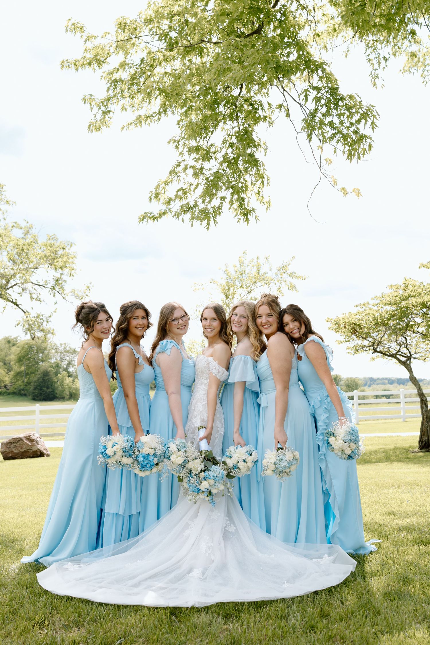 Bride in white gown poses with six bridesmaids in light blue dresses holding floral bouquets outdoors.