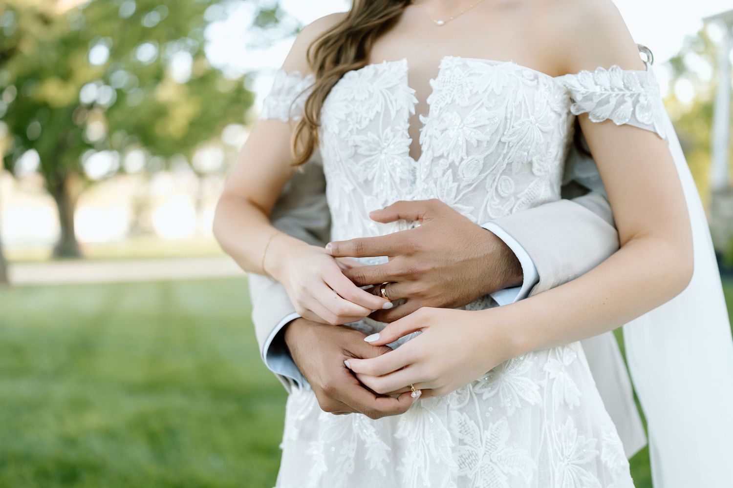 Bride in lace off-shoulder wedding gown embraced by groom, both wearing wedding rings, standing in lush green outdoor setting.