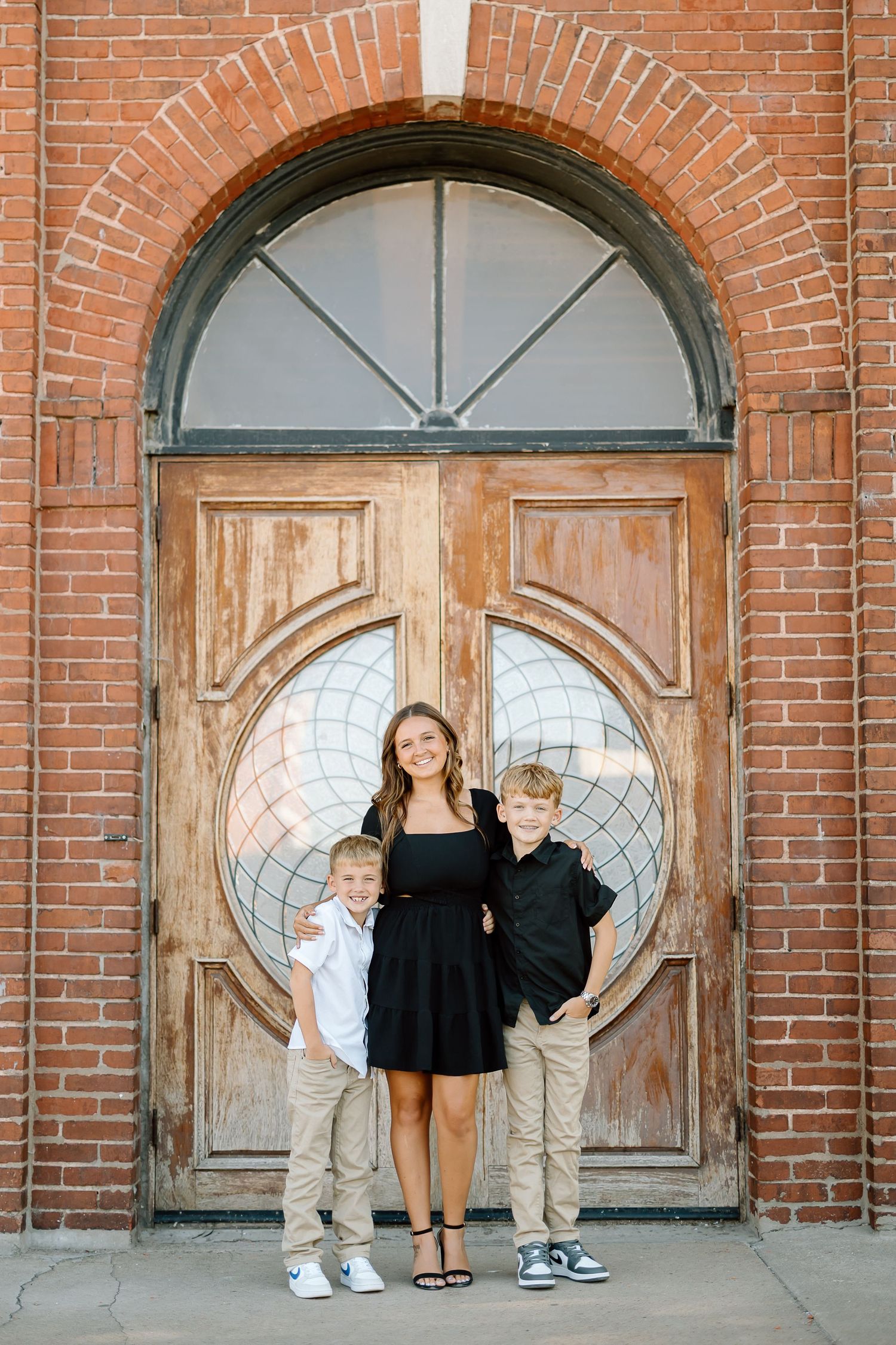 Family stands against rustic brick wall with decorative circular windows and wooden doors in a creative photo series.