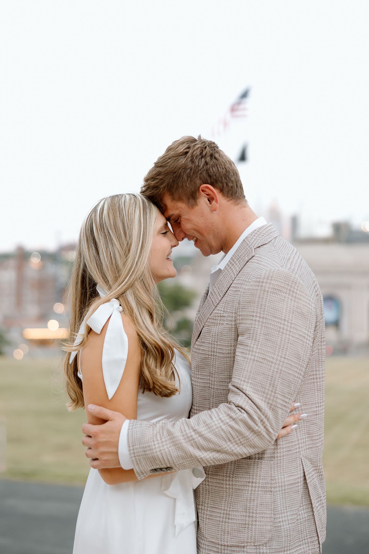A couple shares an intimate moment outdoors, with the person in a white dress and other in a plaid blazer embracing.