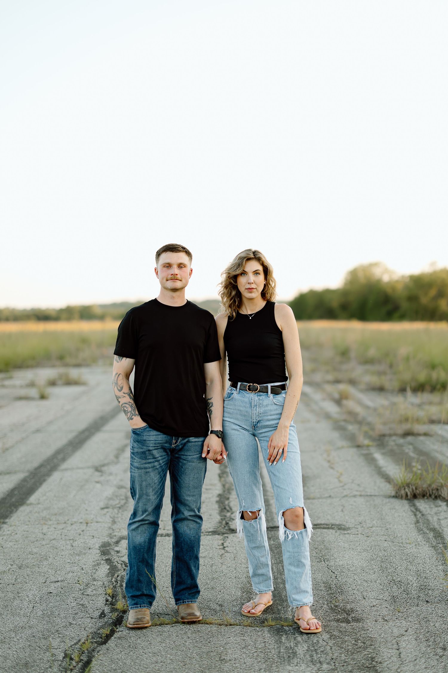 A couple wearing black tops and blue jeans stands together on a rural concrete path at sunset.