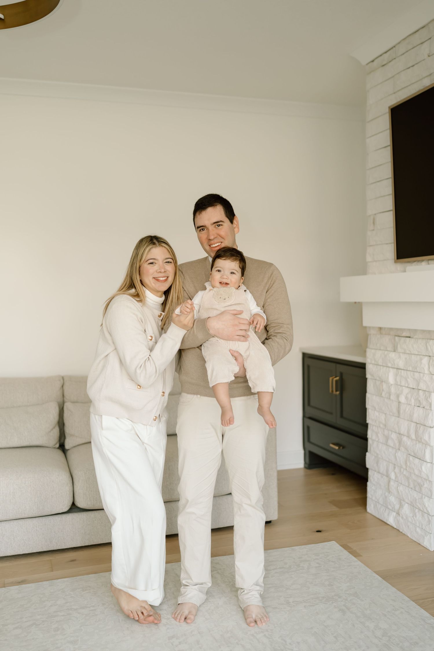 A family in coordinating cream and beige outfits stands barefoot on a gray rug in their modern living room.