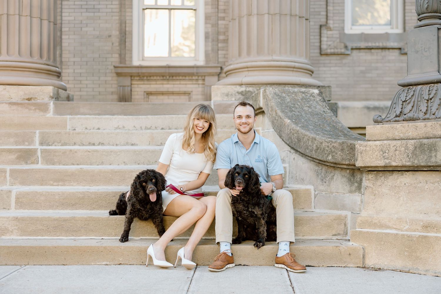 A couple sits on grand stone steps with their two black dogs at a historic building entrance.