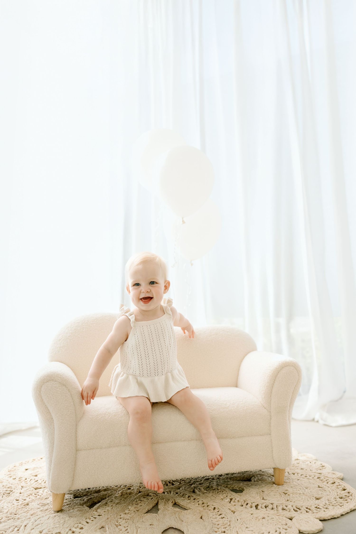 Baby in white dress sits and smiles on elegant cream-colored chaise lounge in bright sunlit room.